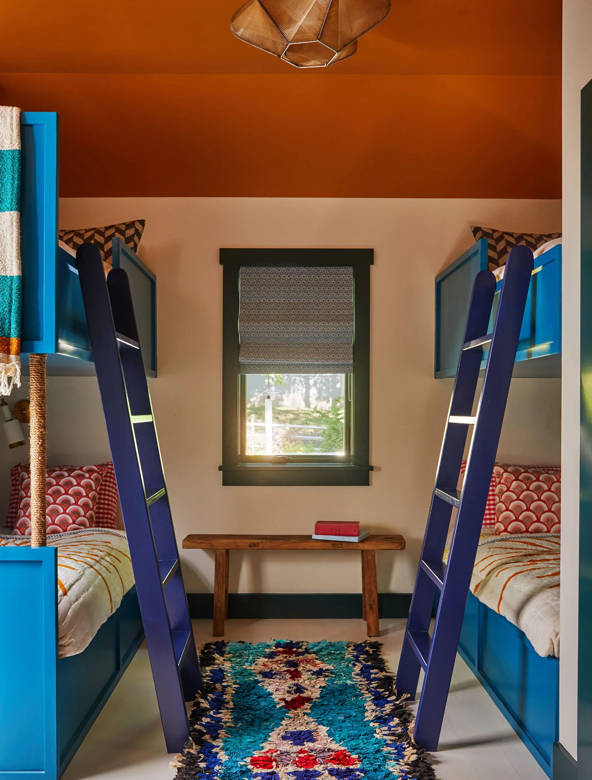 Symmetrical kids bedroom with blue built-in bunk beds flanking a wooden bench and a central window below a terracotta painted ceiling.