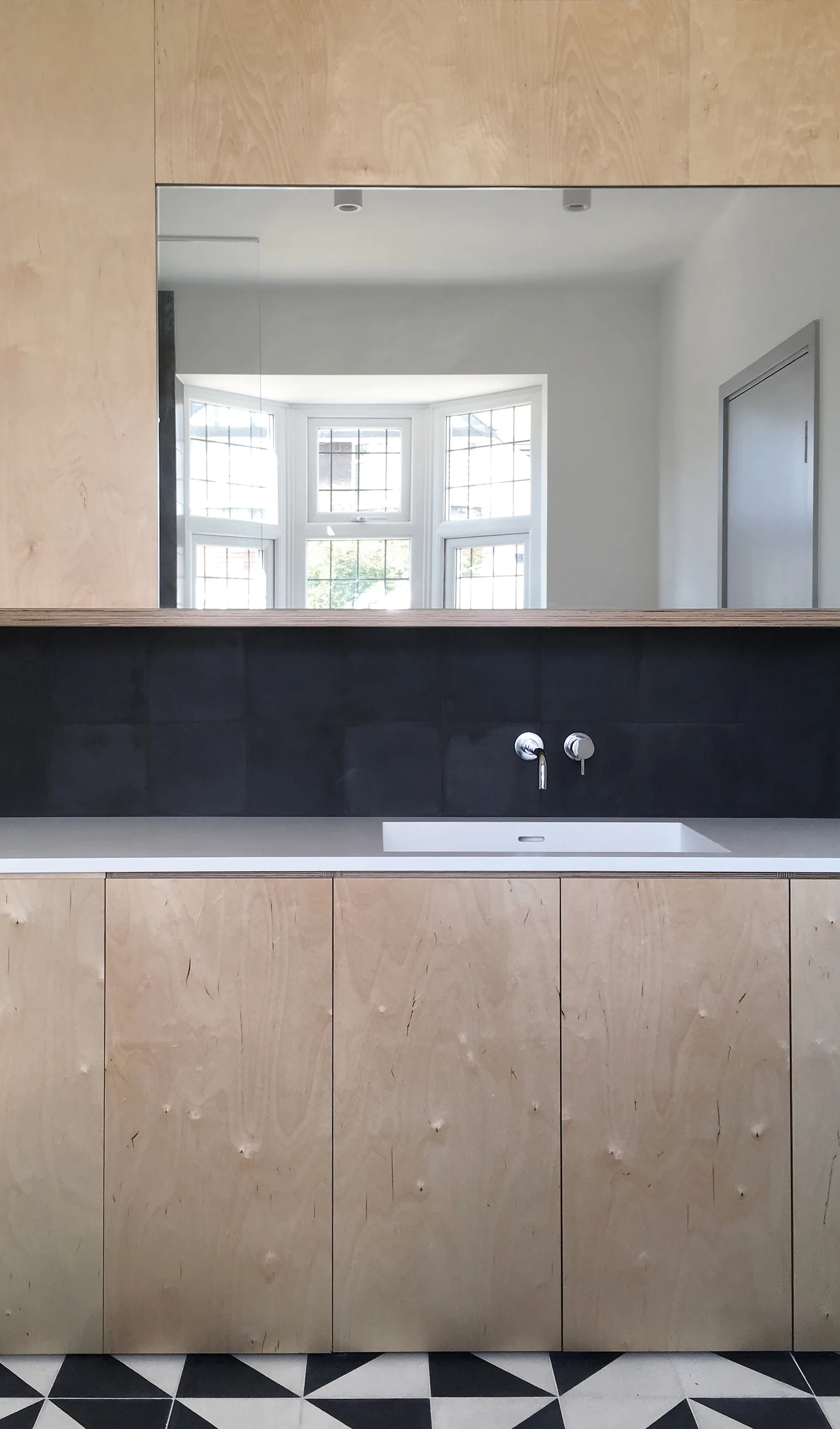A modern kitchen sink area features flat-front plywood cabinets, a matte black tiled backsplash, and a geometric tiled floor.