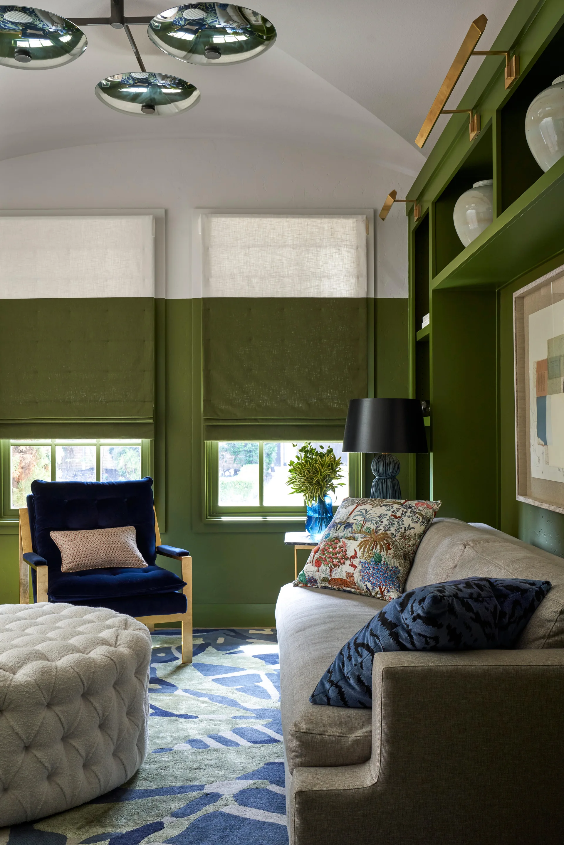 A forest green living room features a gray sofa, a blue velvet armchair, color-blocked roman shades, and built-in shelving topped with brass picture lights.