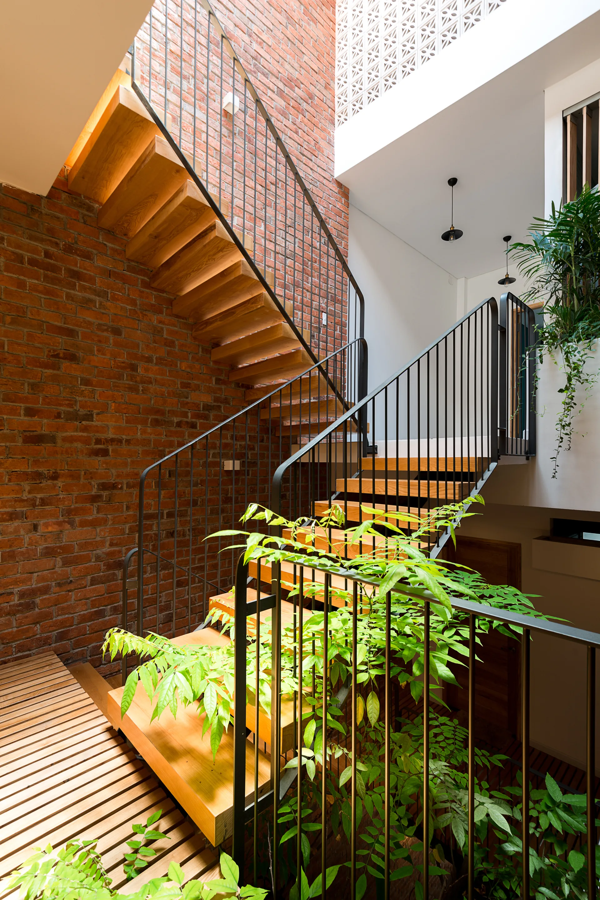 An open riser wooden staircase with black metal railings climbs alongside a tall red brick wall decorated with green hanging plants.