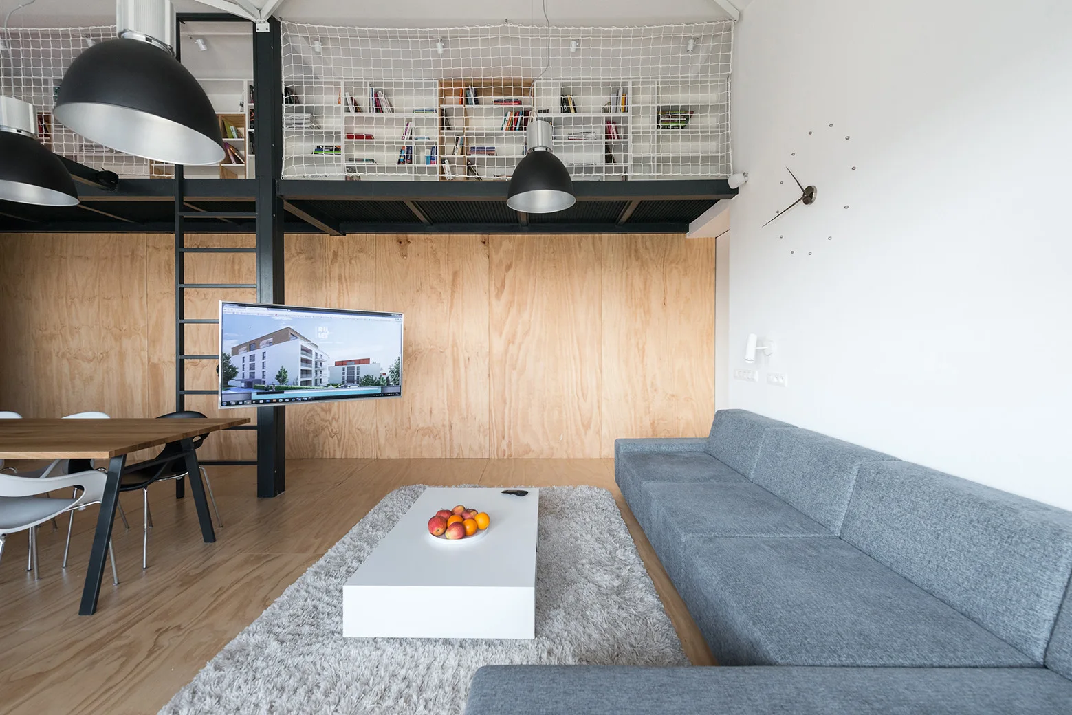 A modern loft living room features a grey sectional sofa, a white coffee table, and a mezzanine level with white netting above a plywood paneled wall.