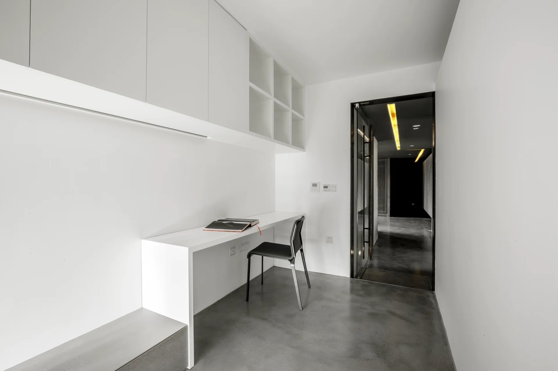 A minimalist home office features a white built-in desk with upper cabinets, a black chair, and a polished gray concrete floor.