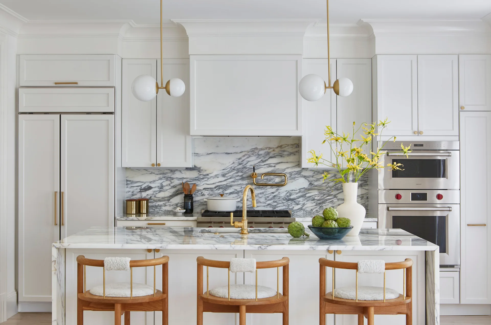 A bright white kitchen featuring a heavily veined marble backsplash, a matching waterfall island, wood bar stools, and gold-toned pendant lights.