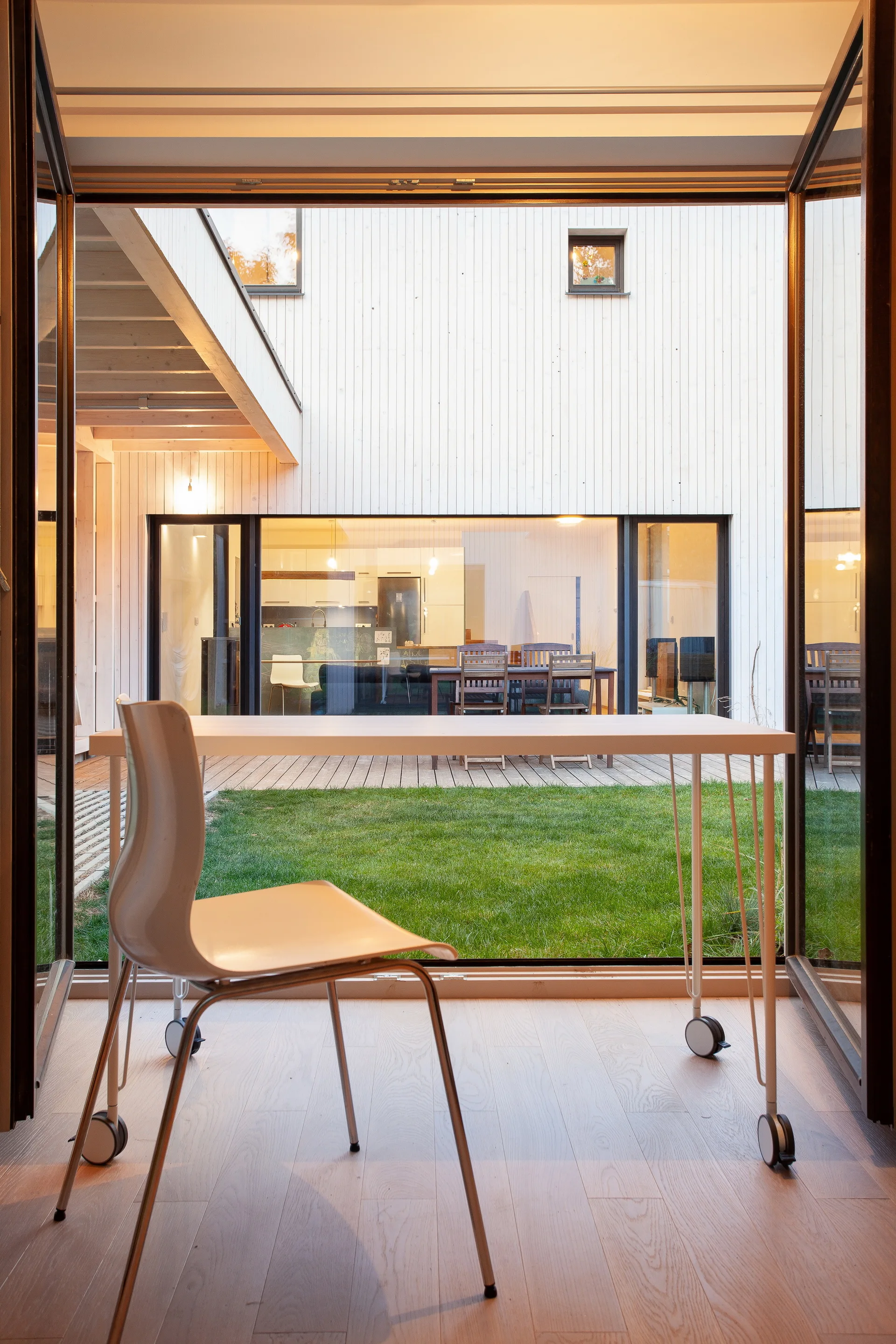 A simple white desk and modern chair stand in front of open folding glass doors that look out onto a grassy courtyard and wood deck.