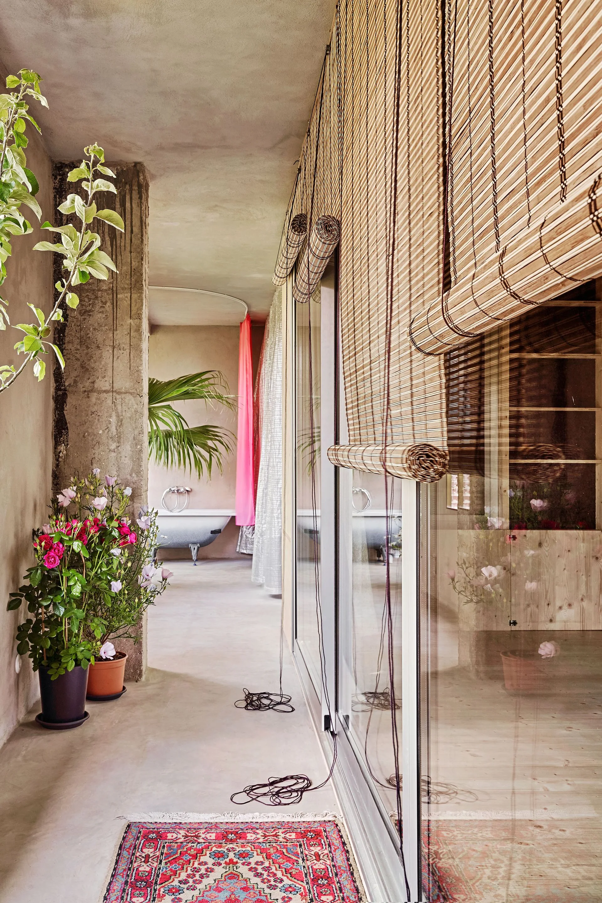 A concrete hallway features bamboo roller shades over a glass sliding door, leading to a bathroom with a freestanding bathtub in the background.