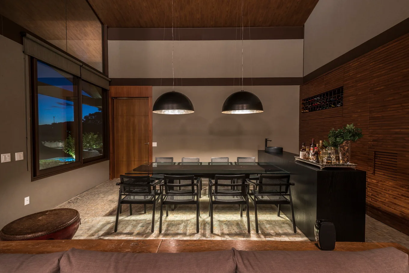 A modern dining room features a long glass-top table, black woven chairs, two oversized dome pendant lights, and textured stone floor pavers.