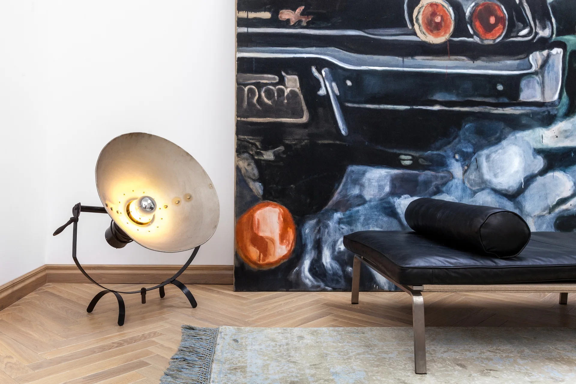 A black leather daybed with a bolster cushion rests on a herringbone wood floor next to an industrial metal spotlight lamp and large leaning art.