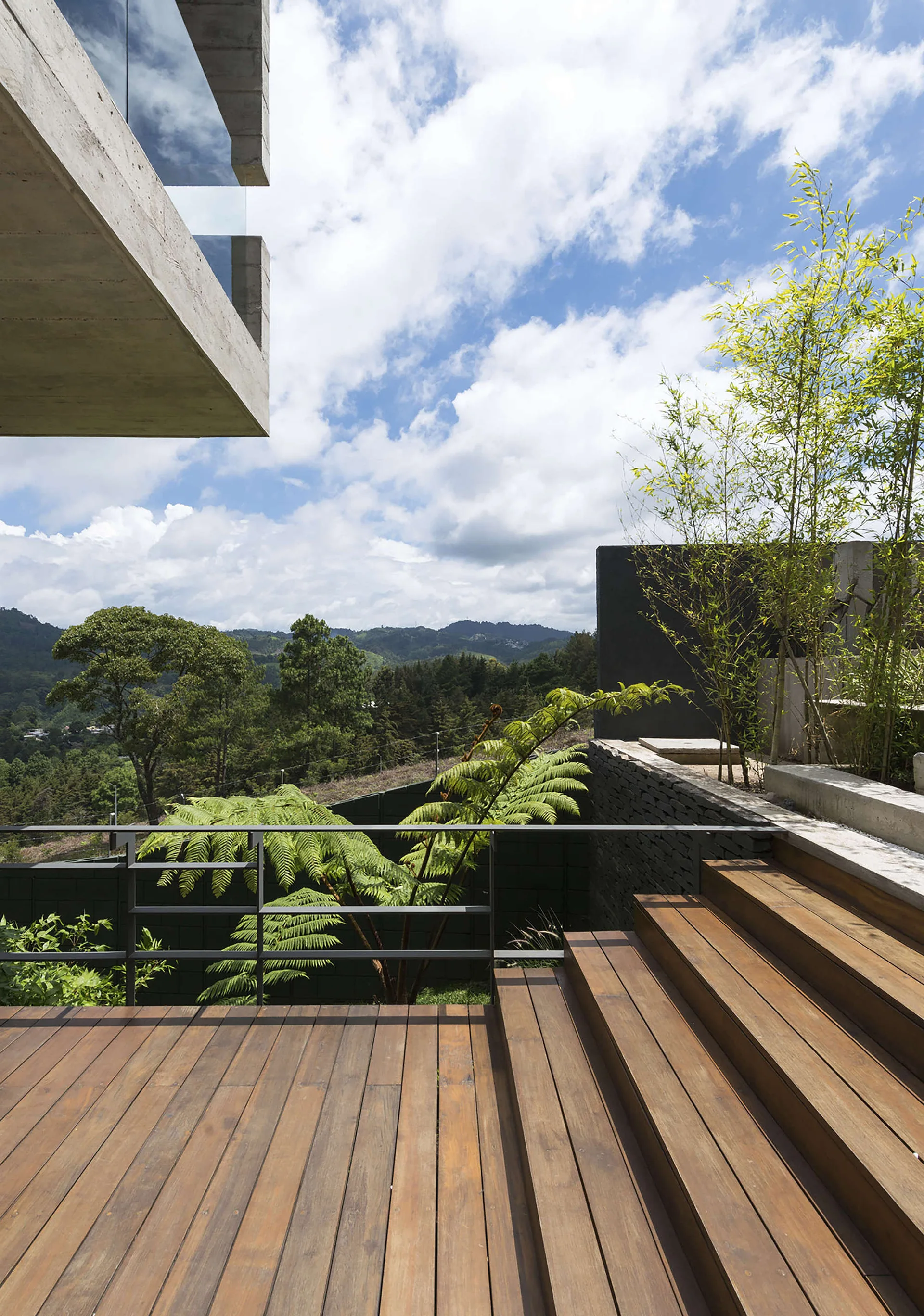 A tiered wood terrace deck features wide timber steps, a black railing, and a raw concrete overhang overlooking a mountain forest landscape.