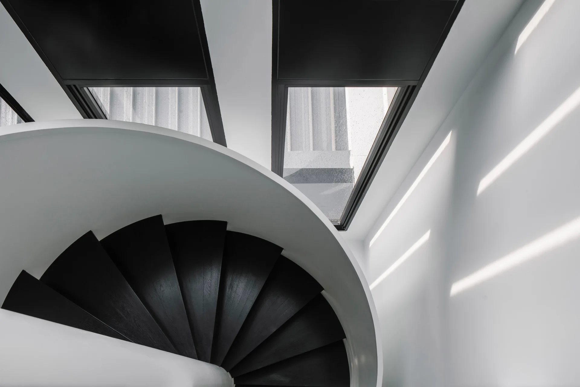 A high-angle view looking down a curved black wooden spiral staircase surrounded by plain white walls with black-framed windows.