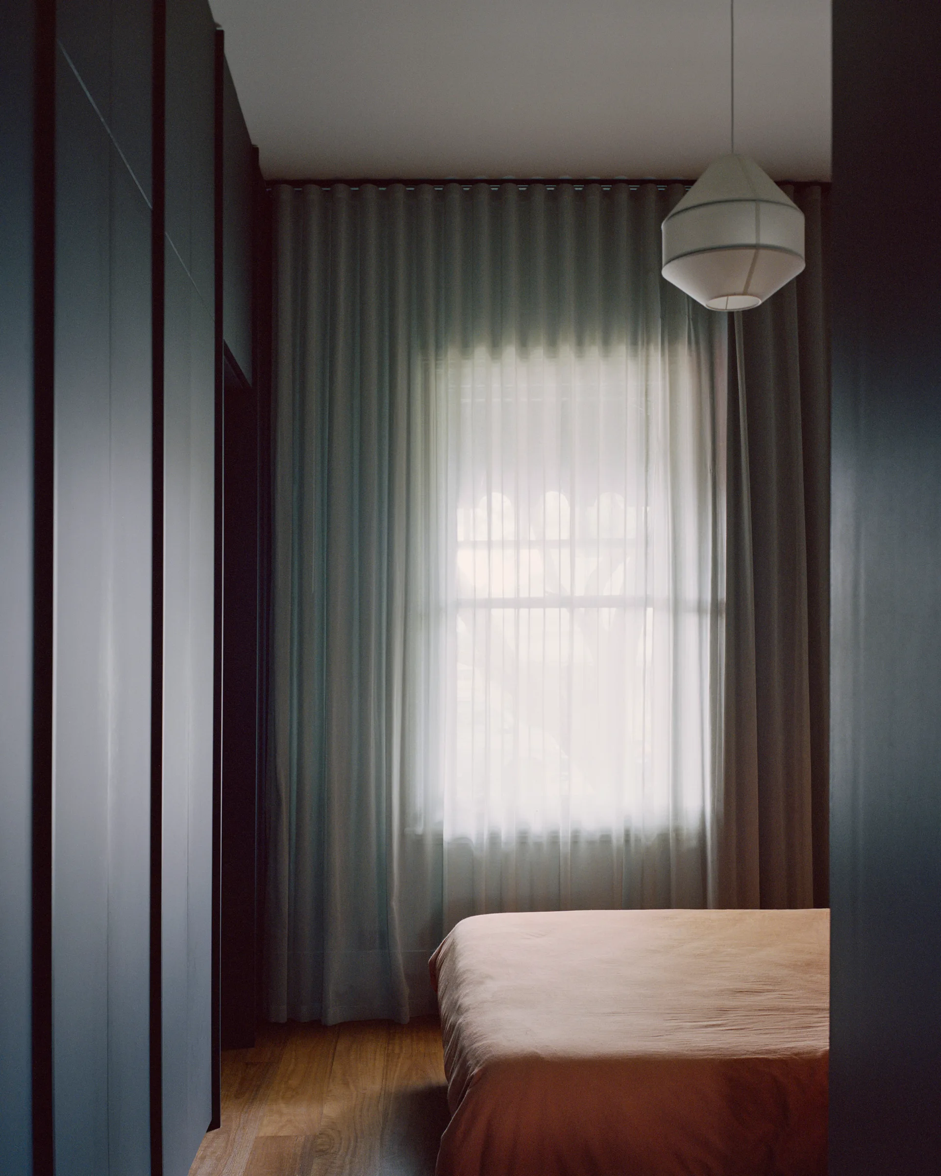 A bedroom features dark charcoal built-in wardrobes, a bed with beige linens, and floor-to-ceiling sheer curtains filtering light from a window.