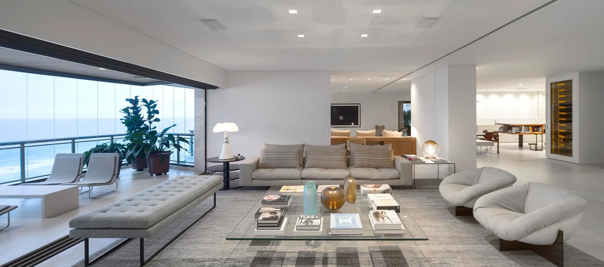 A spacious minimalist living room features a beige sofa, glass coffee table, and tufted bench, opening to a balcony with ocean views.