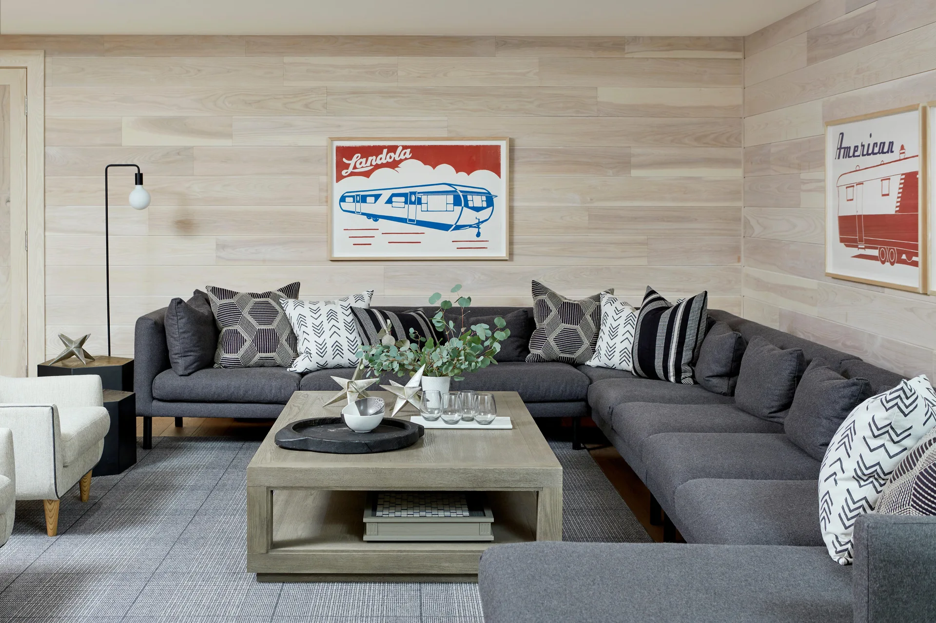 A large charcoal gray sectional sofa sits on a patterned rug in a room with light wood paneled walls and retro framed art.