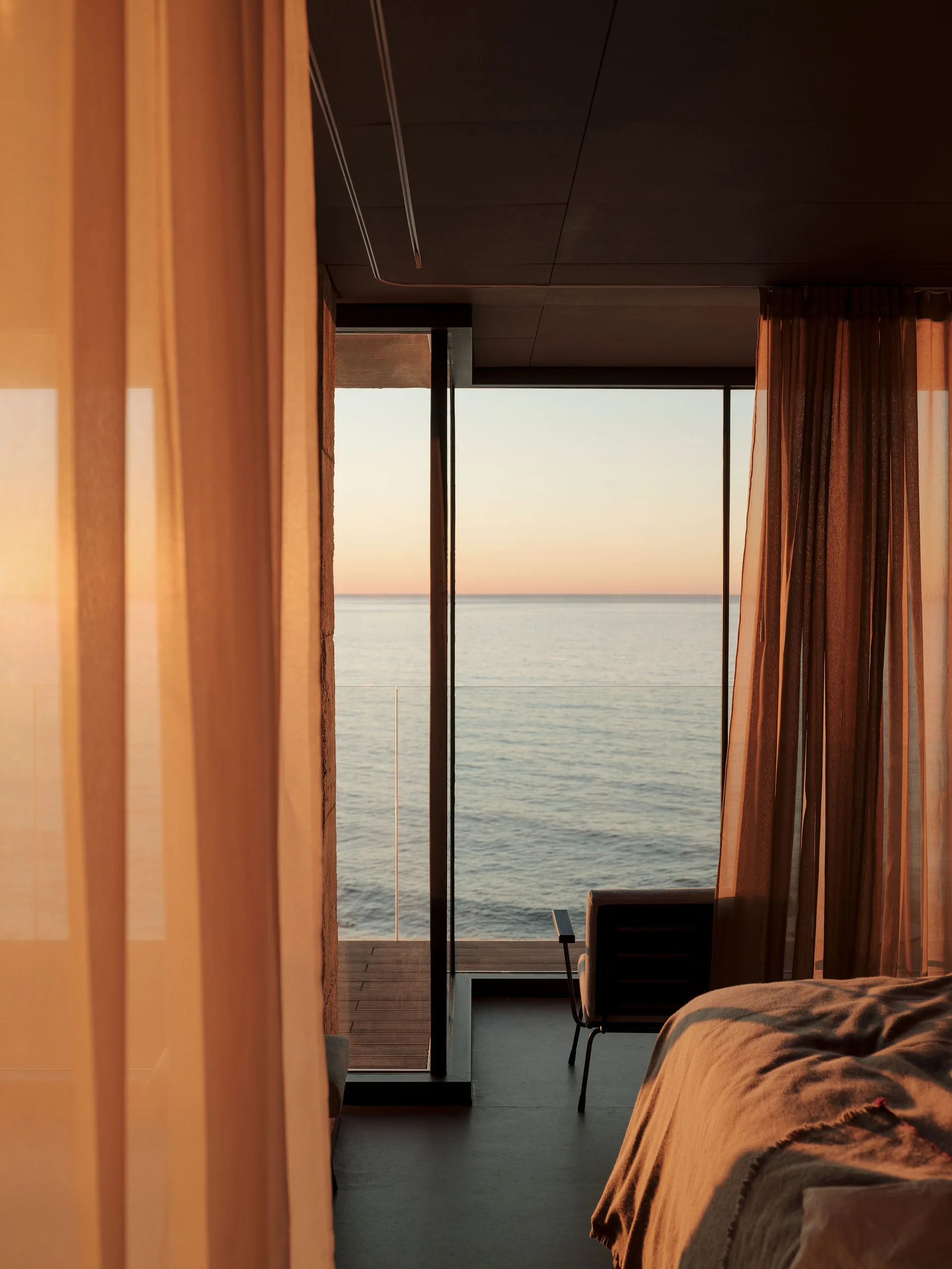 Sheer warm-toned curtains frame a minimalist bedroom window overlooking the ocean at sunset with a simple armchair nearby.