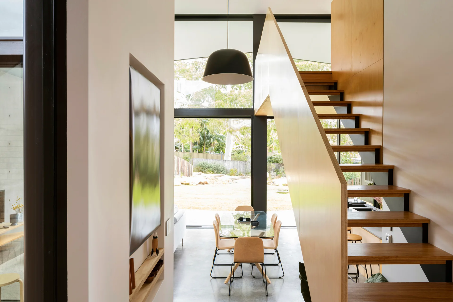 A modern interior features an open wooden staircase, a glass-top dining table, and a large window looking out onto a green yard.