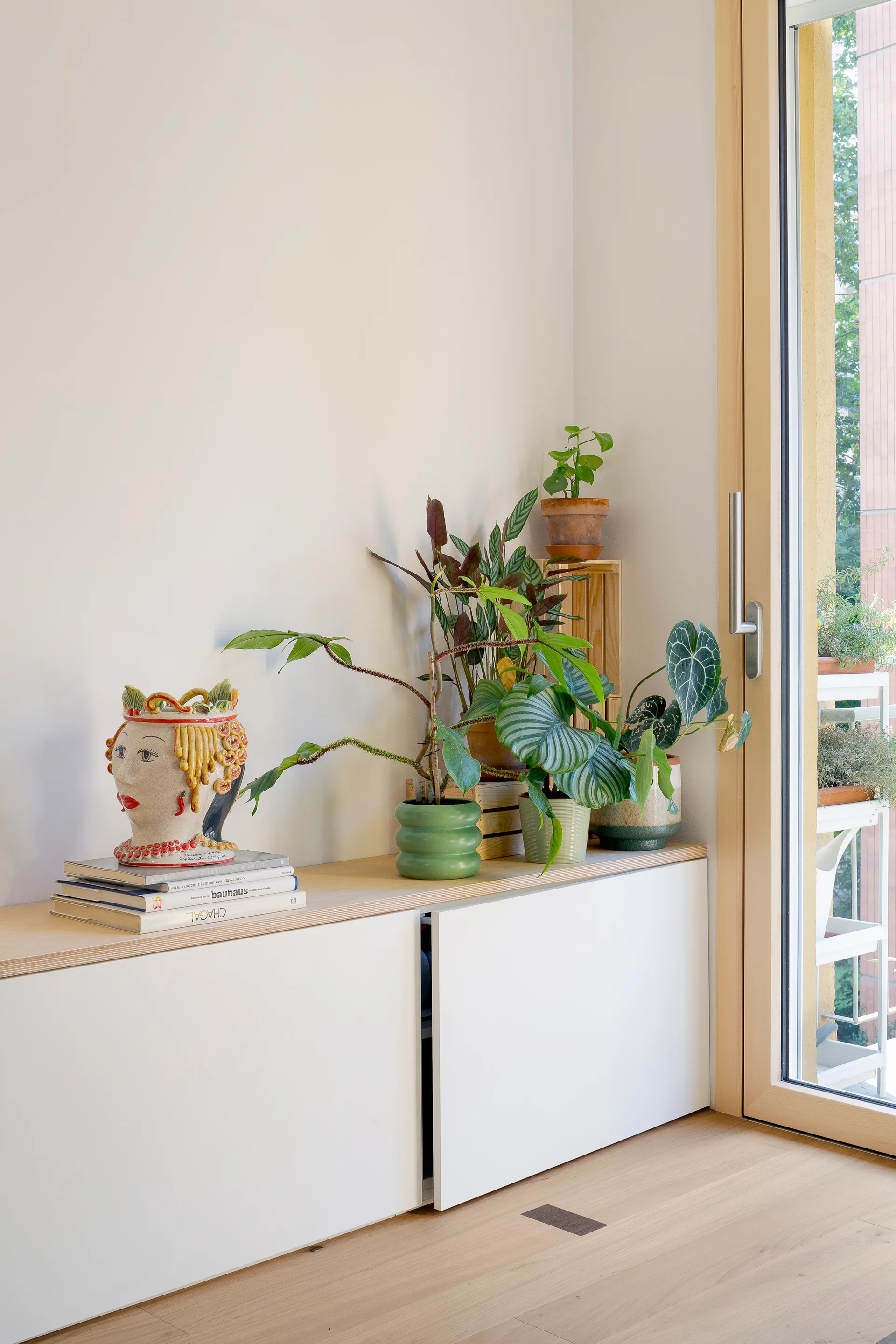 A low white cabinet with a light wood top displays a stack of books topped with a ceramic head vase alongside several potted houseplants next to a window.