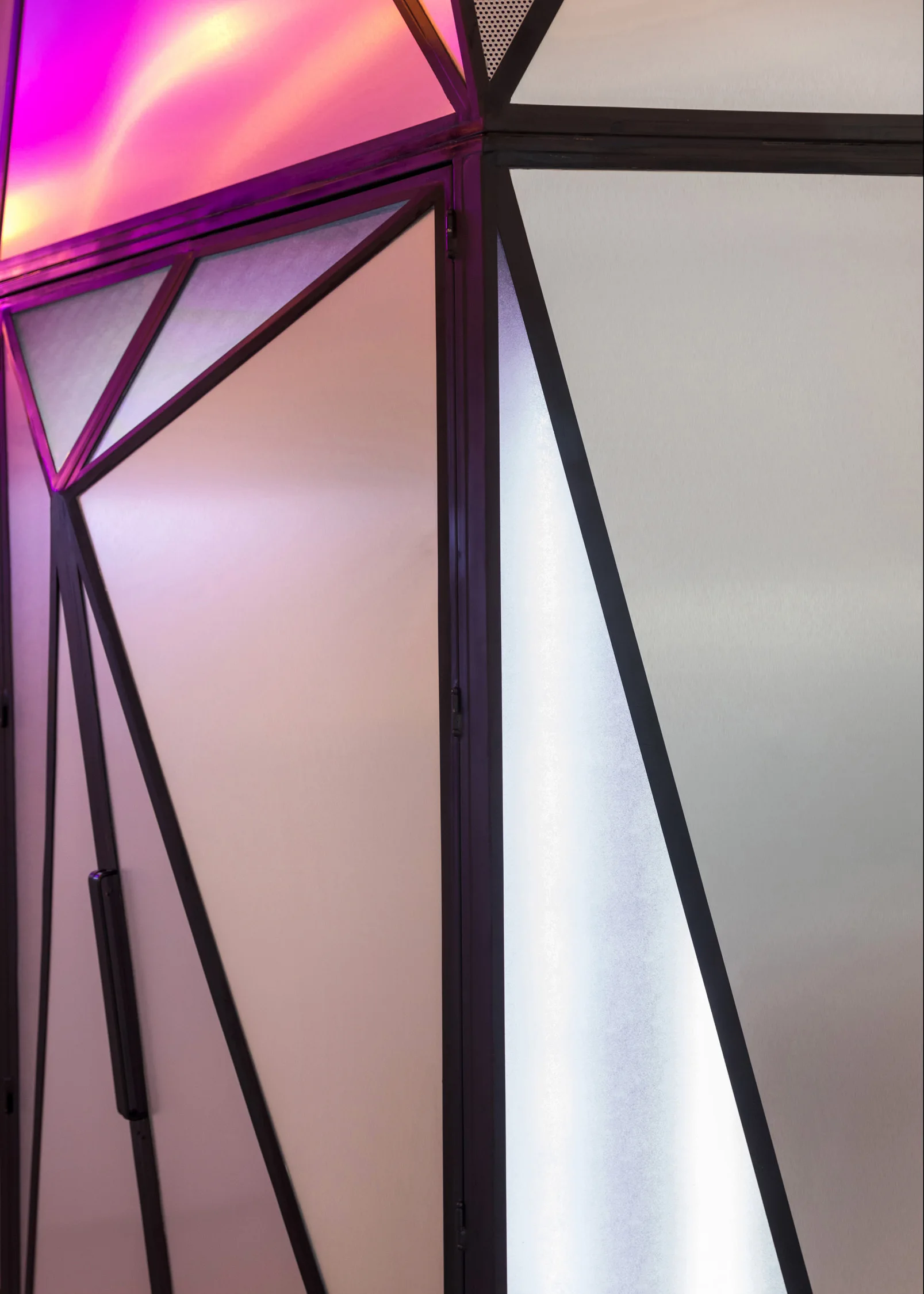 A close-up of a geometric wall partition made of dark metal frames and frosted glass panels illuminated by pink, purple, and white LED lighting.