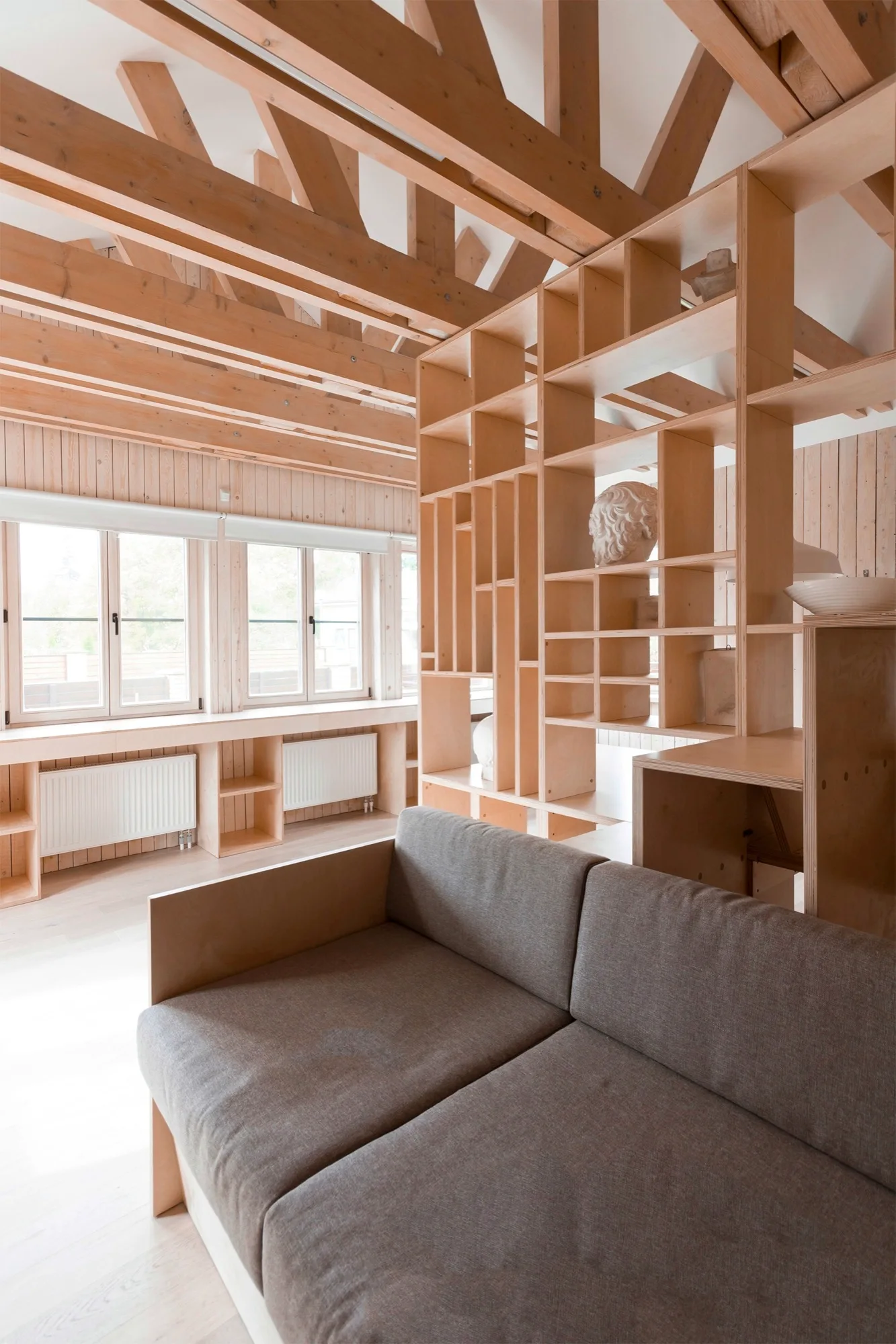 A grey upholstered sofa sits in a room featuring a large plywood shelving partition, wood paneled walls, and exposed timber ceiling trusses.