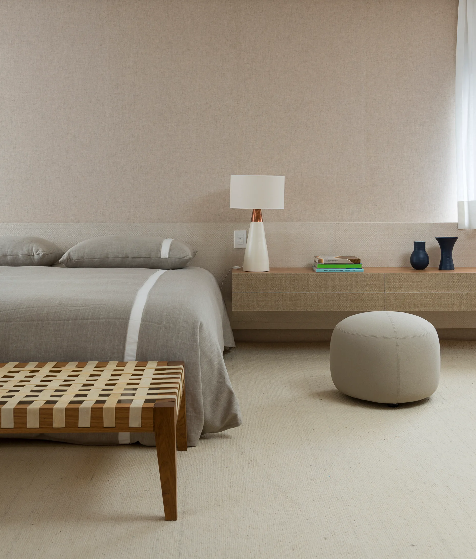 A minimalist bedroom with a grey linen bed, a woven leather-strap bench, a floating wooden nightstand, a table lamp, and a round grey ottoman.