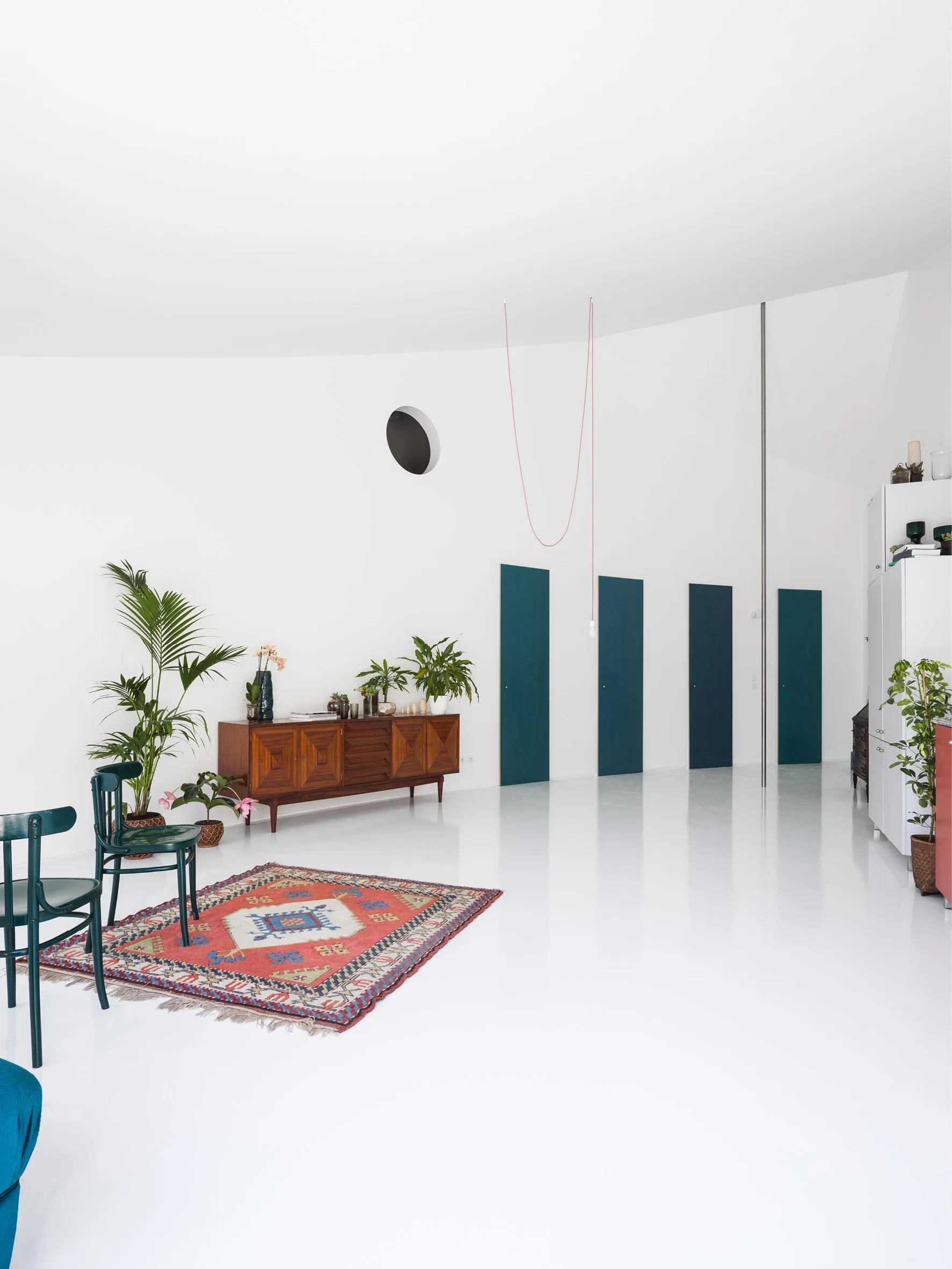 A spacious white room features a curved wall with four teal doors, a mid-century wooden sideboard, and a patterned rug on a glossy white floor.