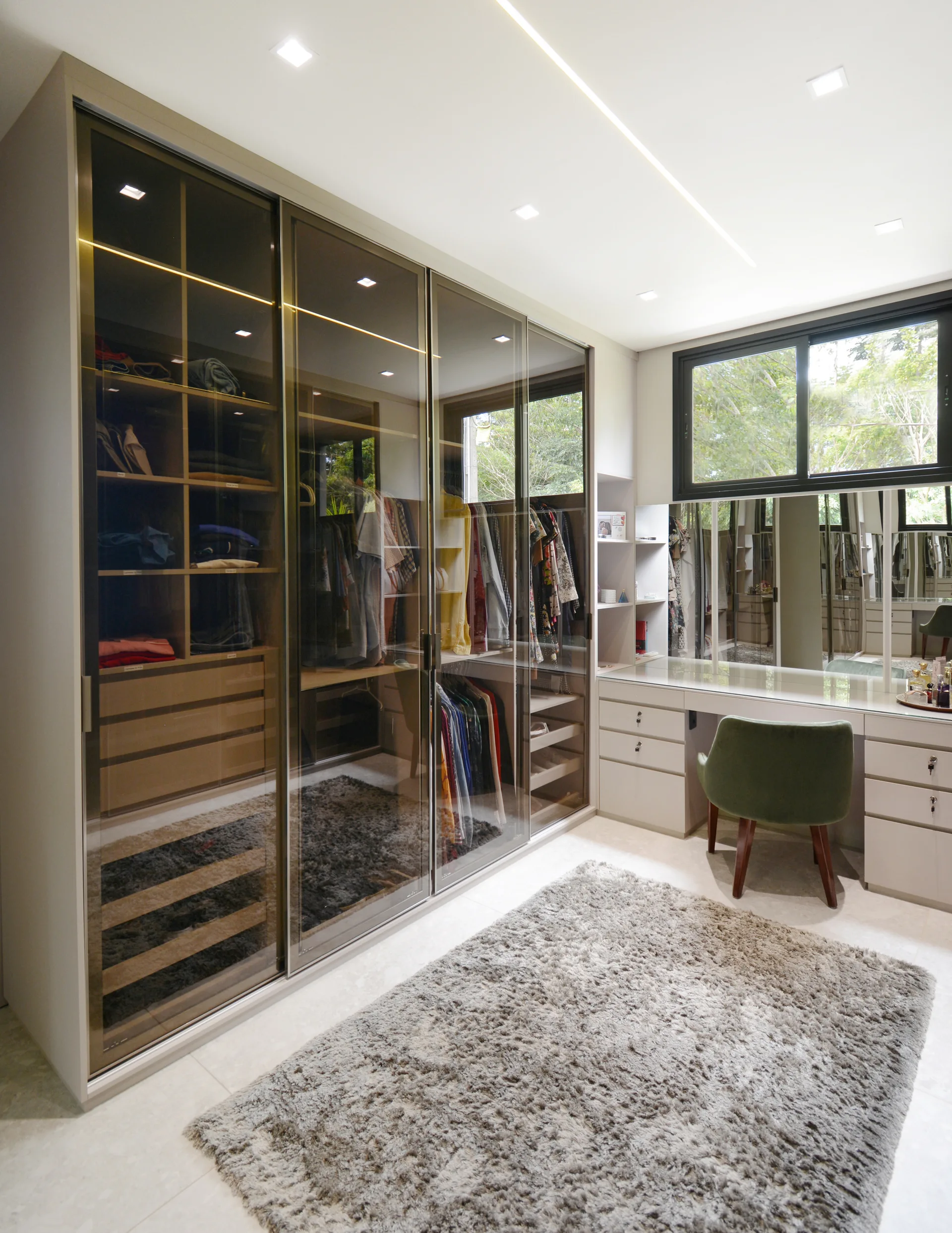 A walk-in dressing room featuring a glass-front wardrobe with internal lighting, a built-in vanity table, a green chair, and a grey shag rug.