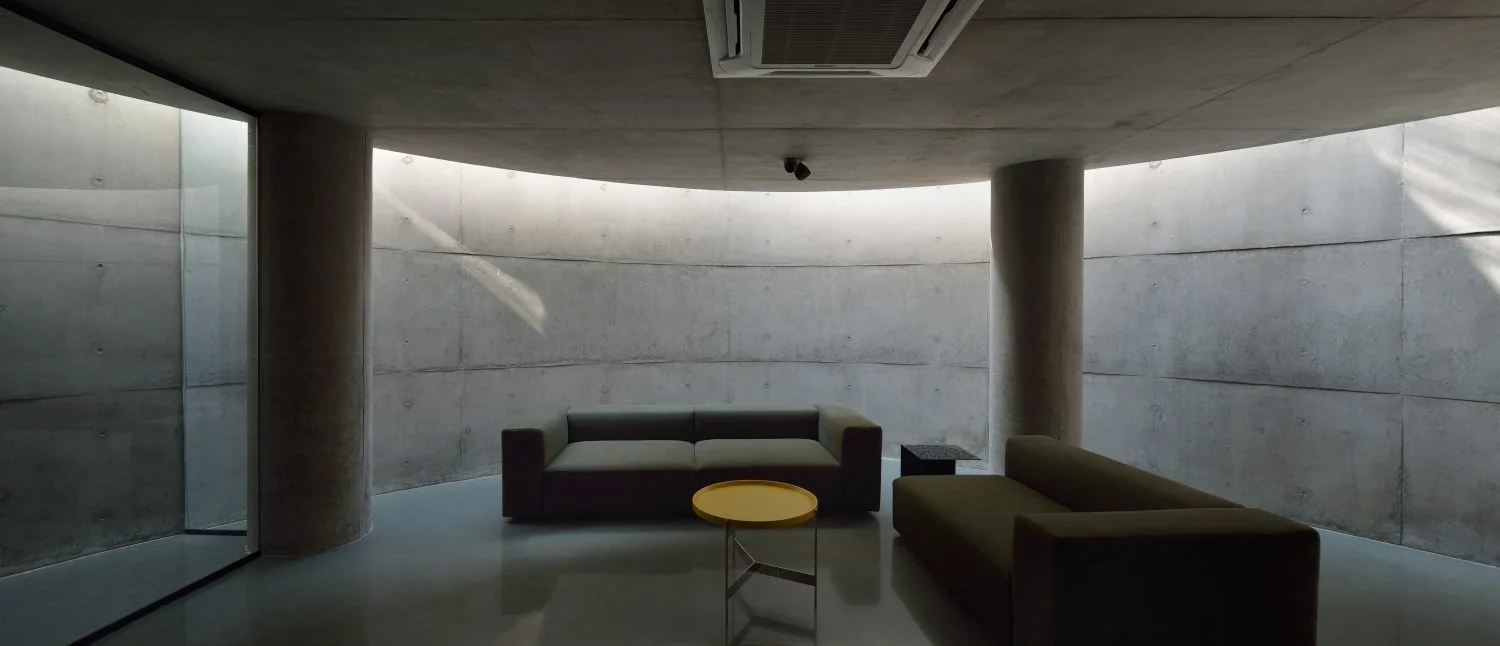 A minimalist living room featuring two low-profile green sofas and a small yellow table set against a curved concrete wall with overhead light.