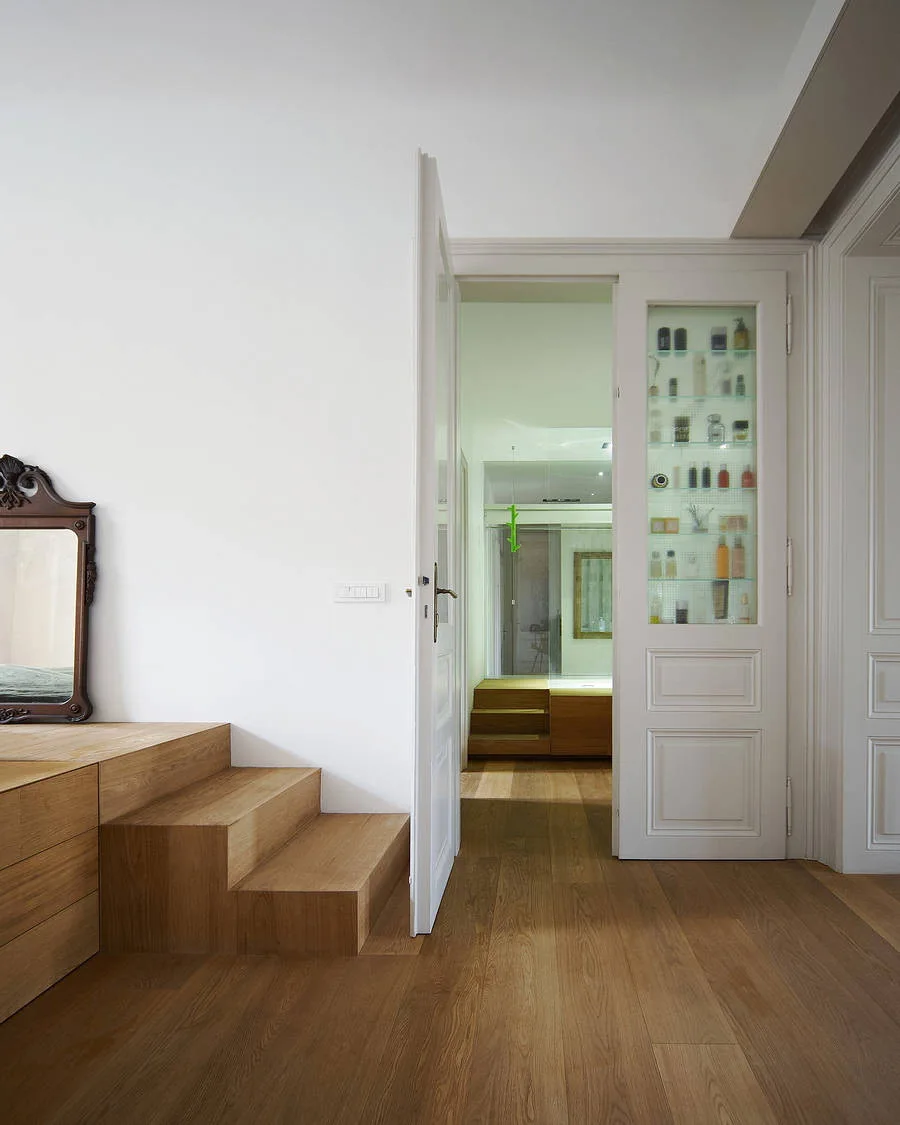 A minimalist white hallway features light oak floors, custom built-in wooden platform steps, and a white door with integrated glass display shelves.
