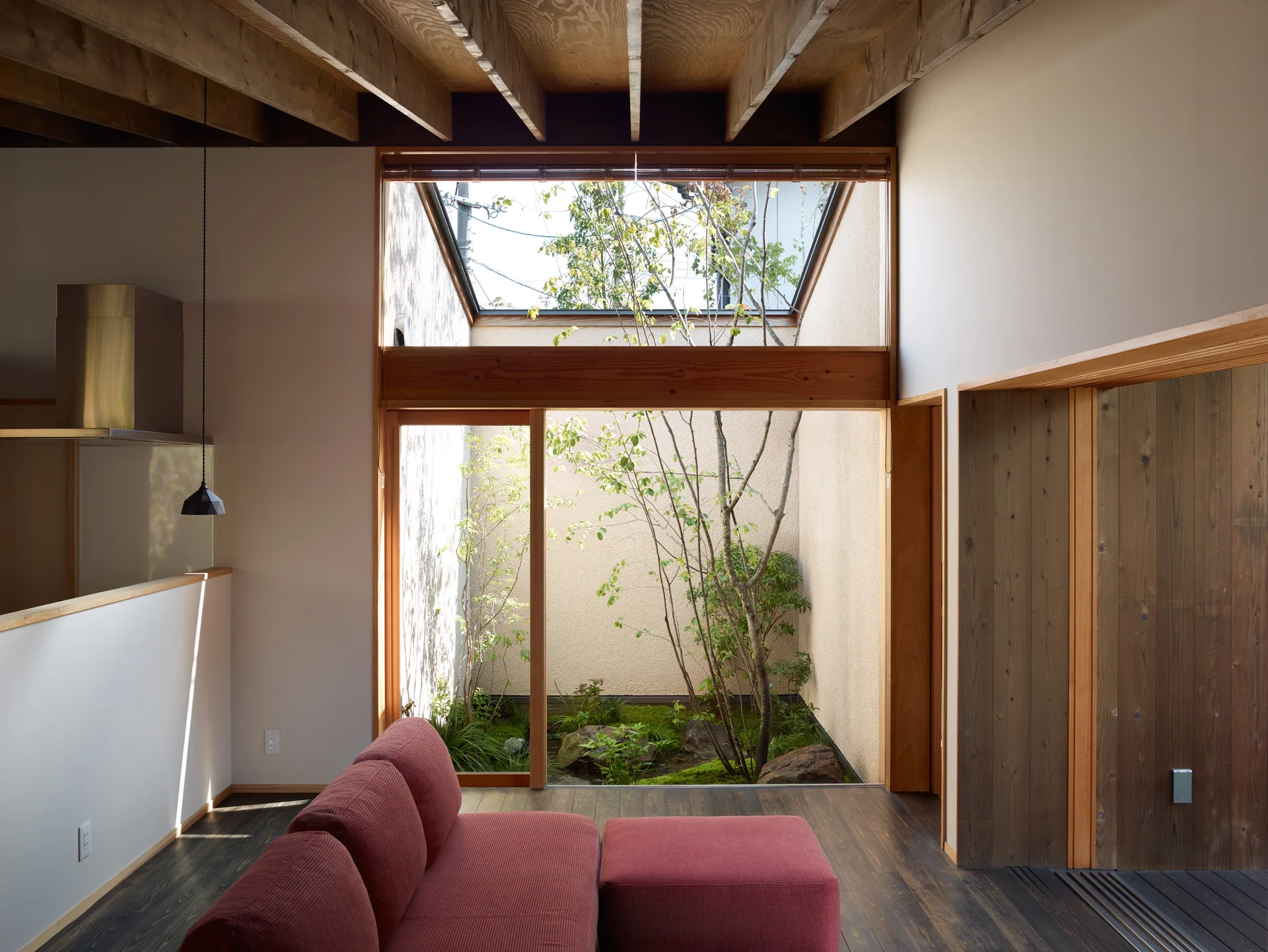 A minimalist living room featuring a red fabric sofa, dark wood floors, exposed ceiling beams, and a large window looking onto a private courtyard.