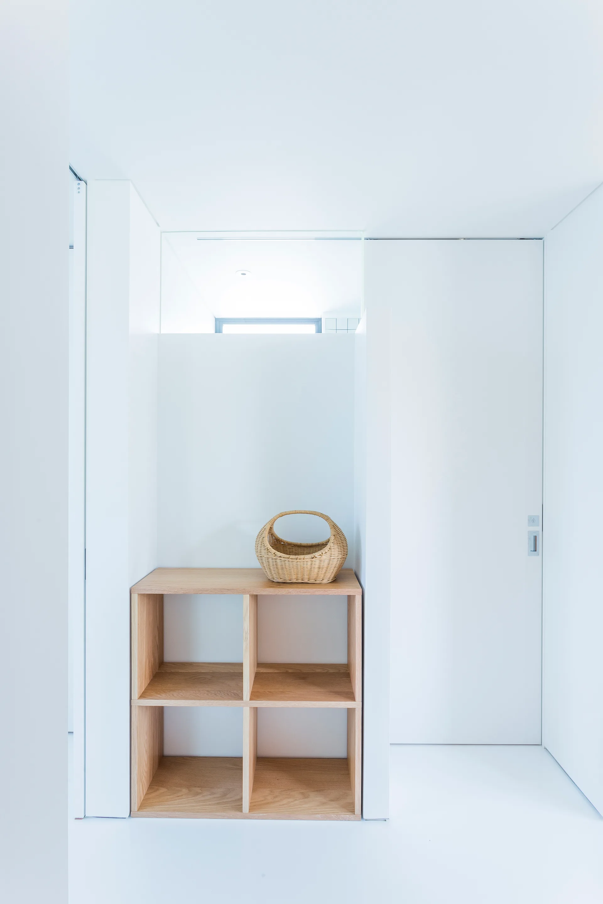 A minimalist white entryway features a light wood cubby shelving unit holding a woven wicker basket next to a white flush door.