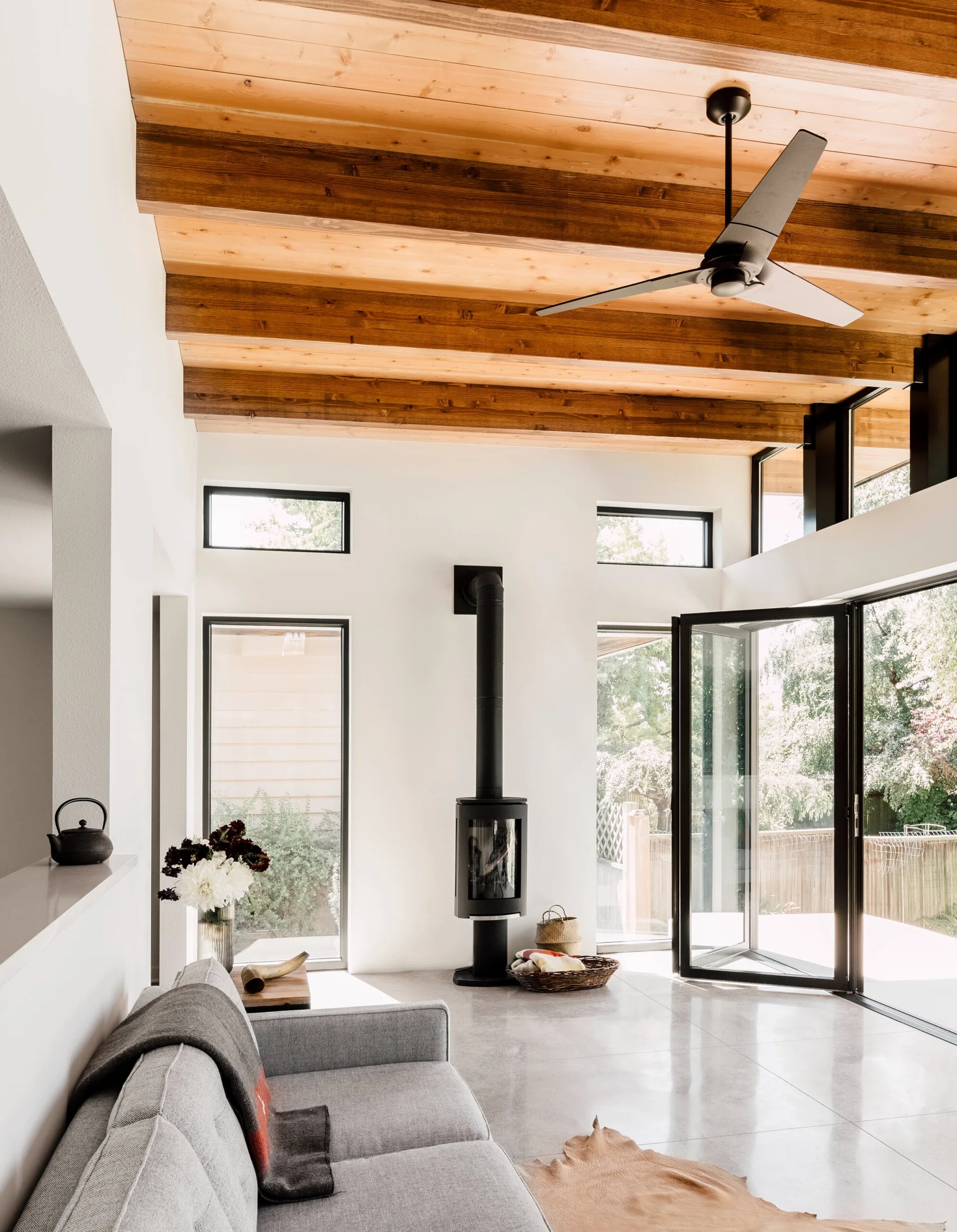 A modern living room featuring a warm wood ceiling with exposed beams, a black cylindrical wood stove, a grey sofa, and folding glass doors.
