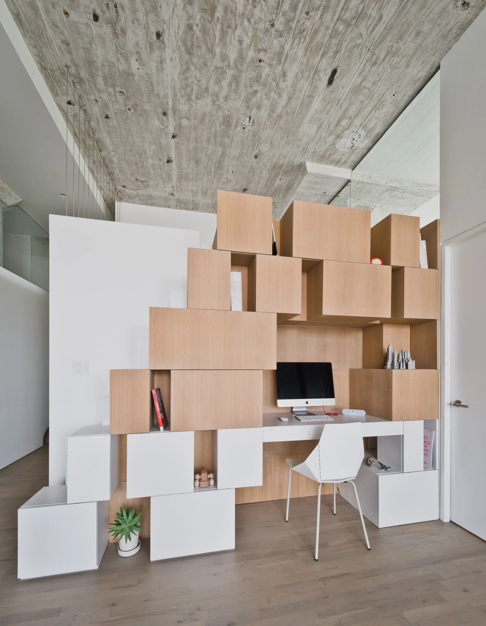 A modern home office featuring a modular stacked wooden shelving unit that incorporates a built-in white desk with a computer and chair.