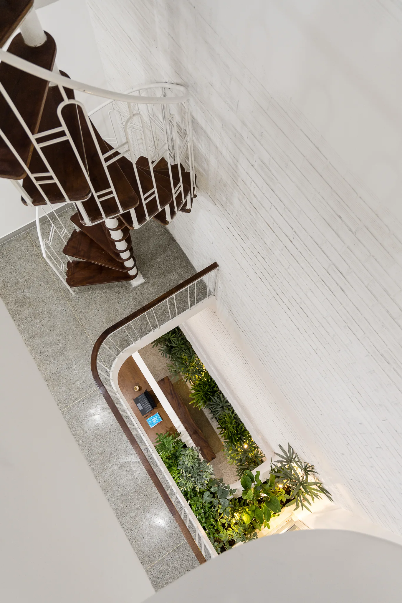 A high-angle view down an atrium featuring a wooden spiral staircase, white brick wall, and indoor plants on the ground floor.