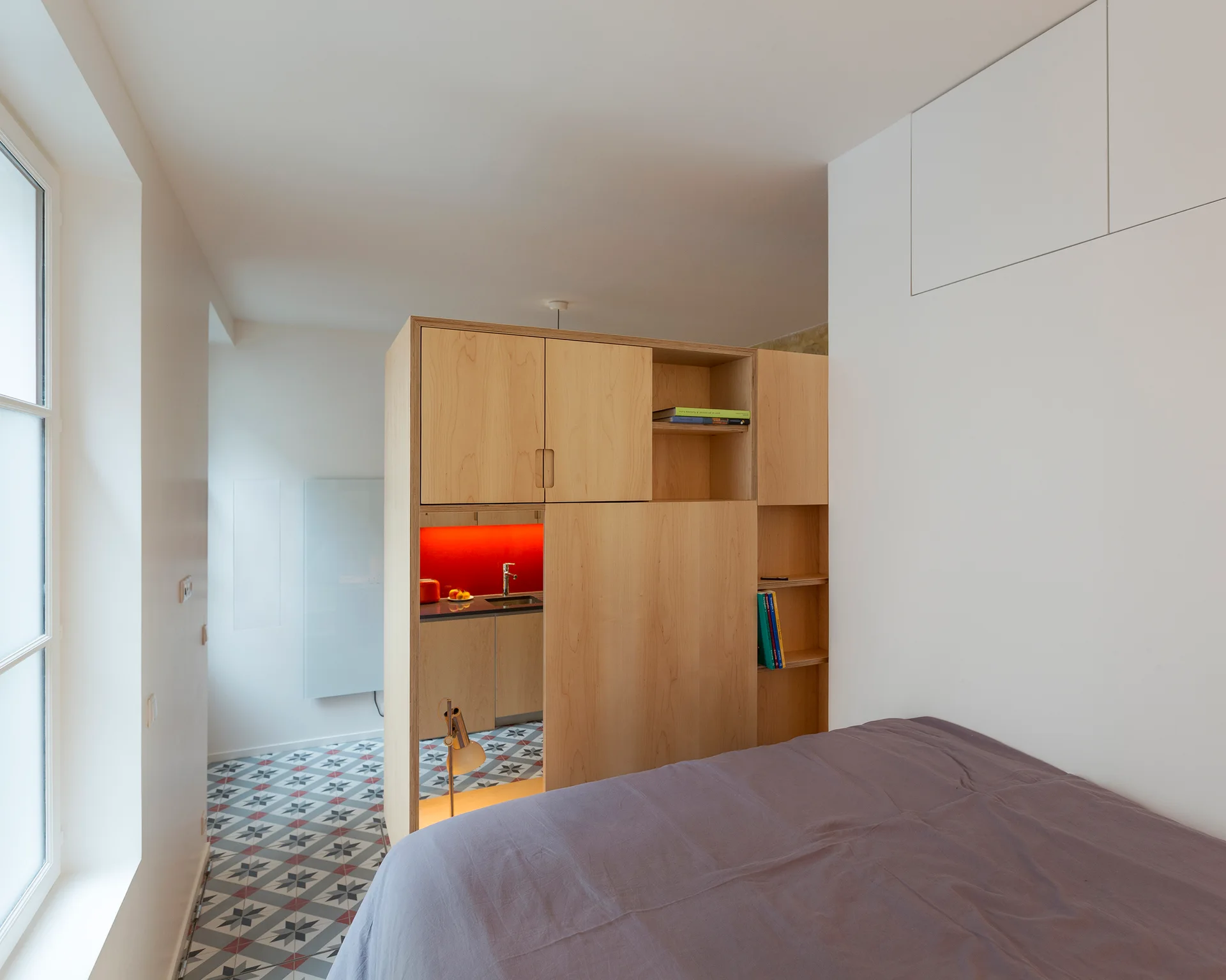 A studio apartment features a bed in the foreground and a light wood partition cabinet dividing the bedroom from a small kitchen with patterned tile floor.