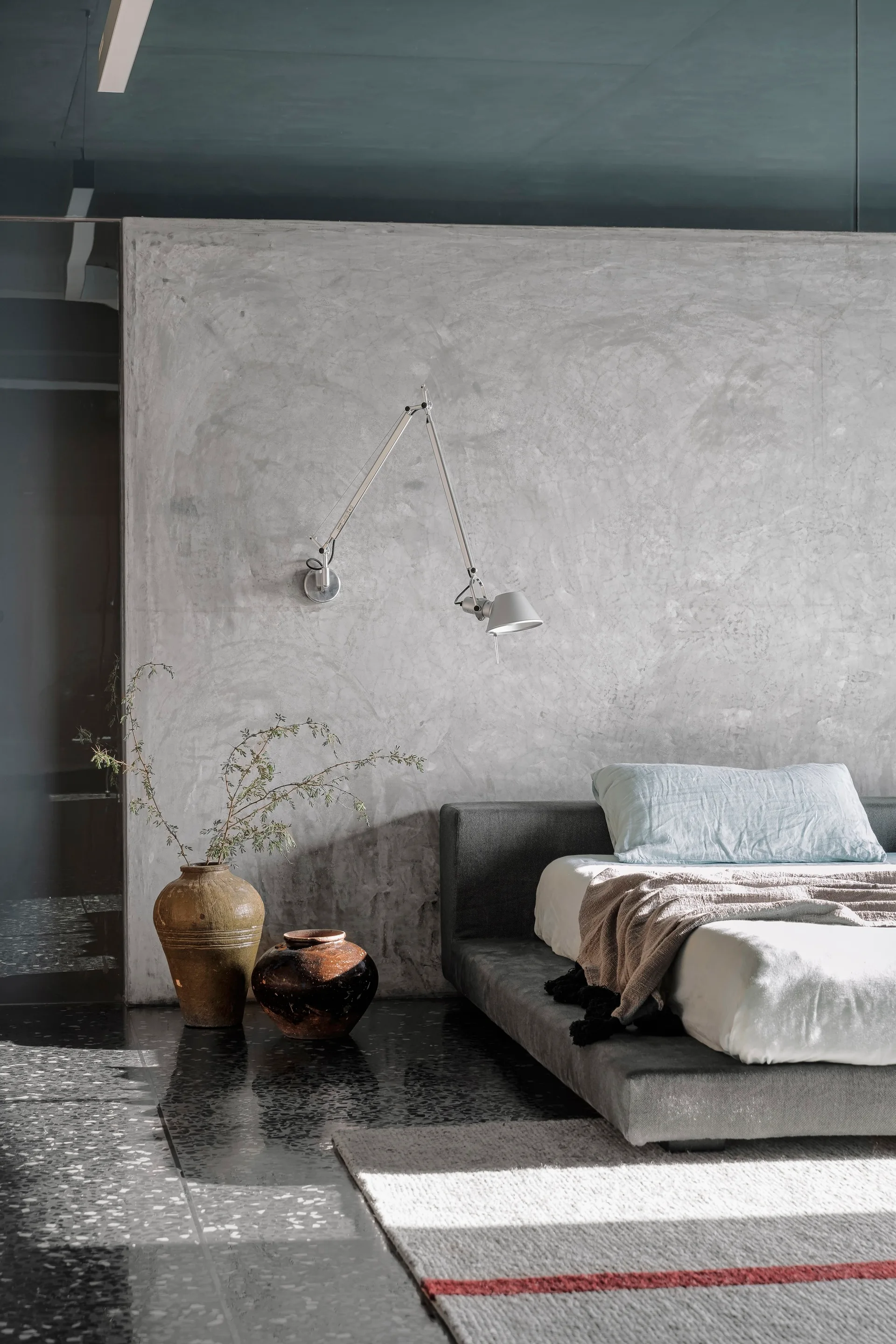 A low-profile grey bed rests against a textured concrete wall next to two floor vases on dark terrazzo flooring.