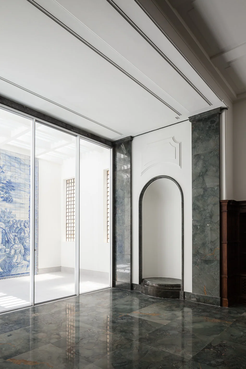 A spacious hallway featuring dark green marble flooring, a recessed arched niche with marble trim, and a large glass partition showing a blue tiled mural.