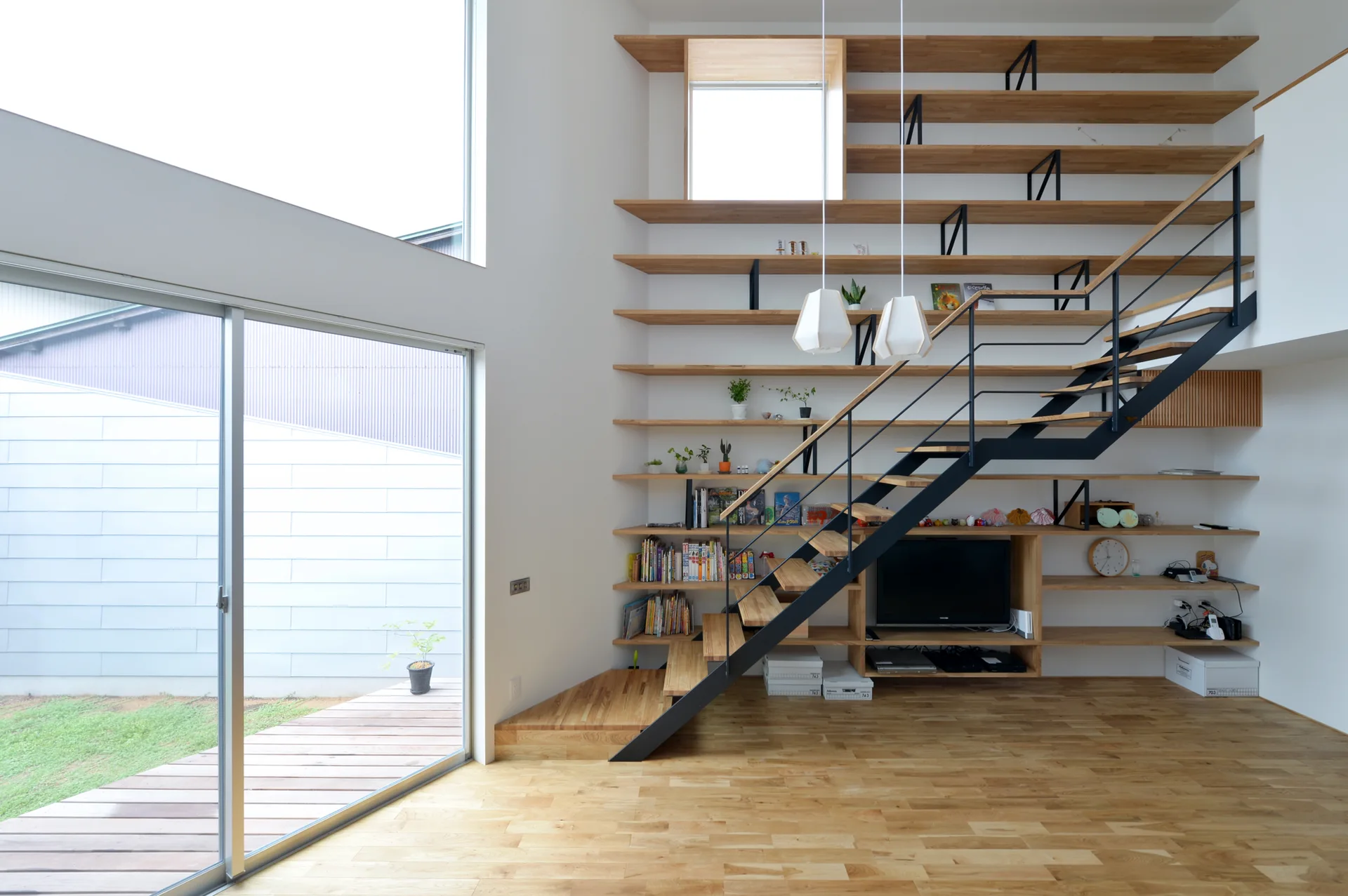 A black metal staircase crosses a large wall-mounted light wood shelving unit in a bright room with hardwood floors and sliding glass doors.