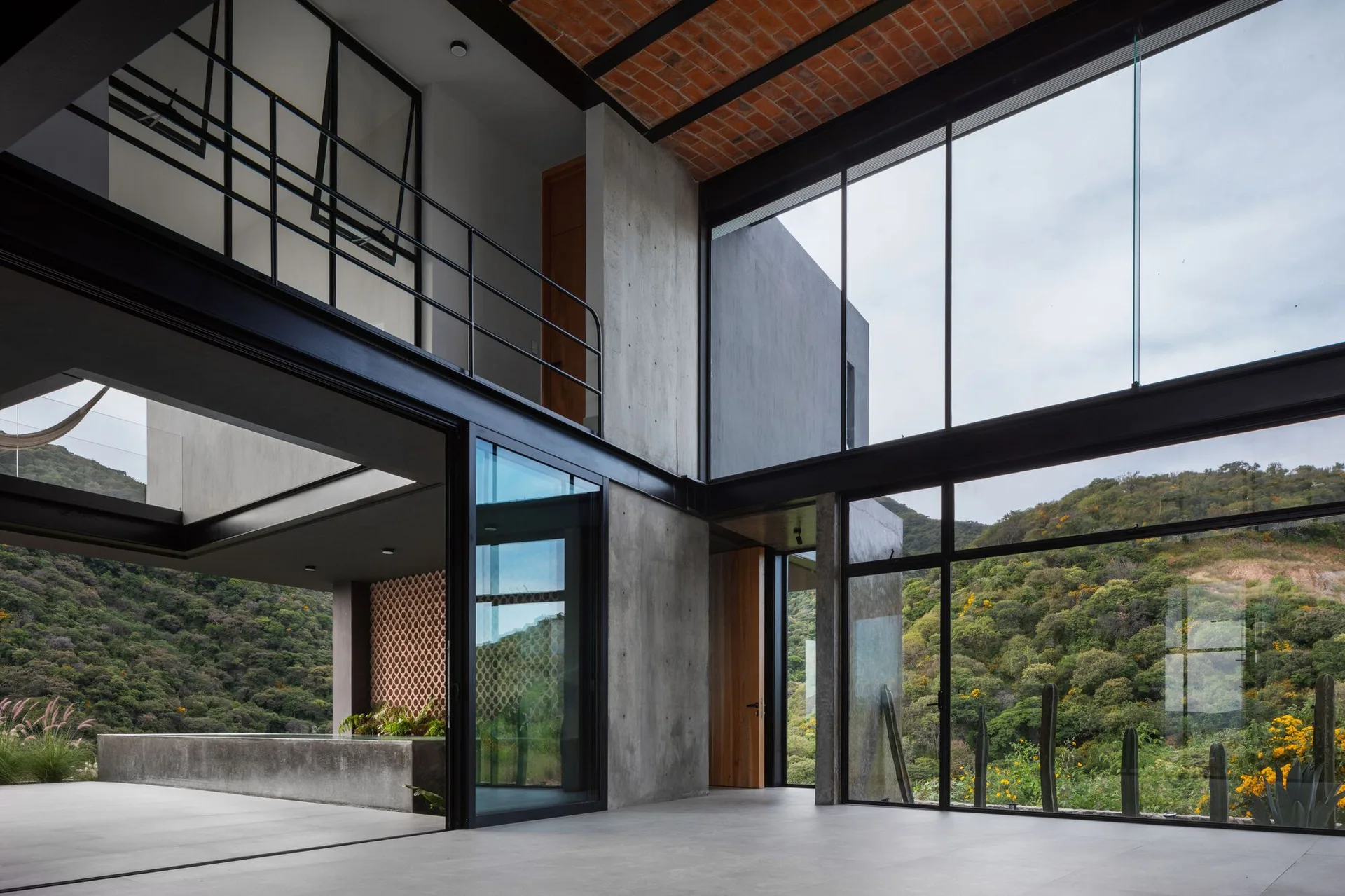 An industrial living area features exposed concrete walls, black steel beams, large glass walls, and a red brick ceiling overlooking a hillside.