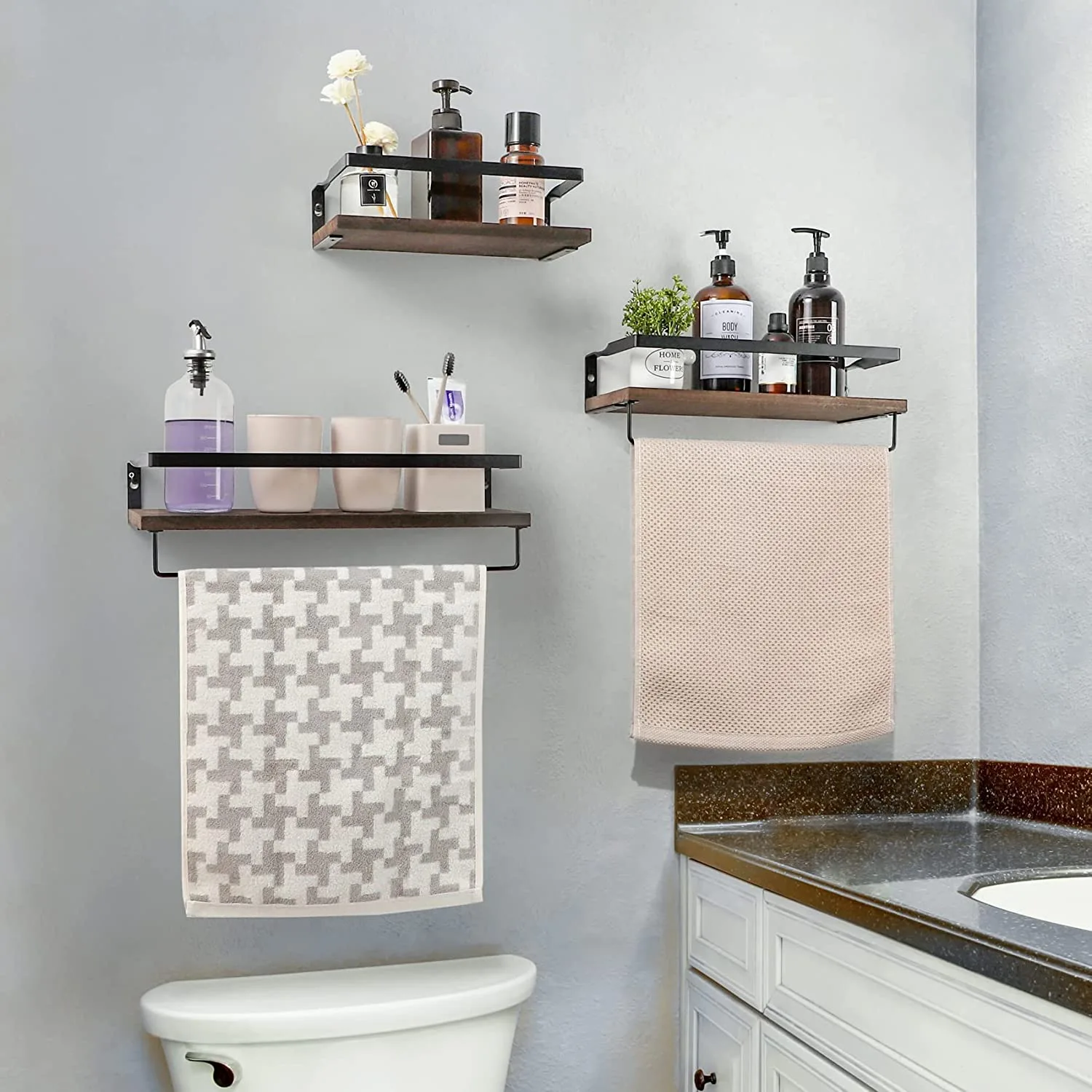 Three staggered dark wood floating shelves with matte black metal rails are mounted on a light gray bathroom wall.