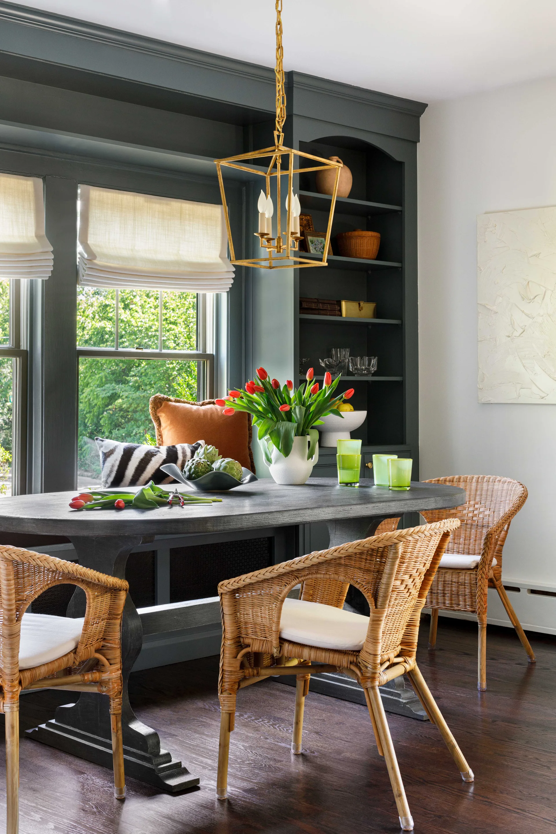 A dark wood dining table surrounded by woven rattan chairs sits near a window flanked by charcoal-painted built-in shelving.