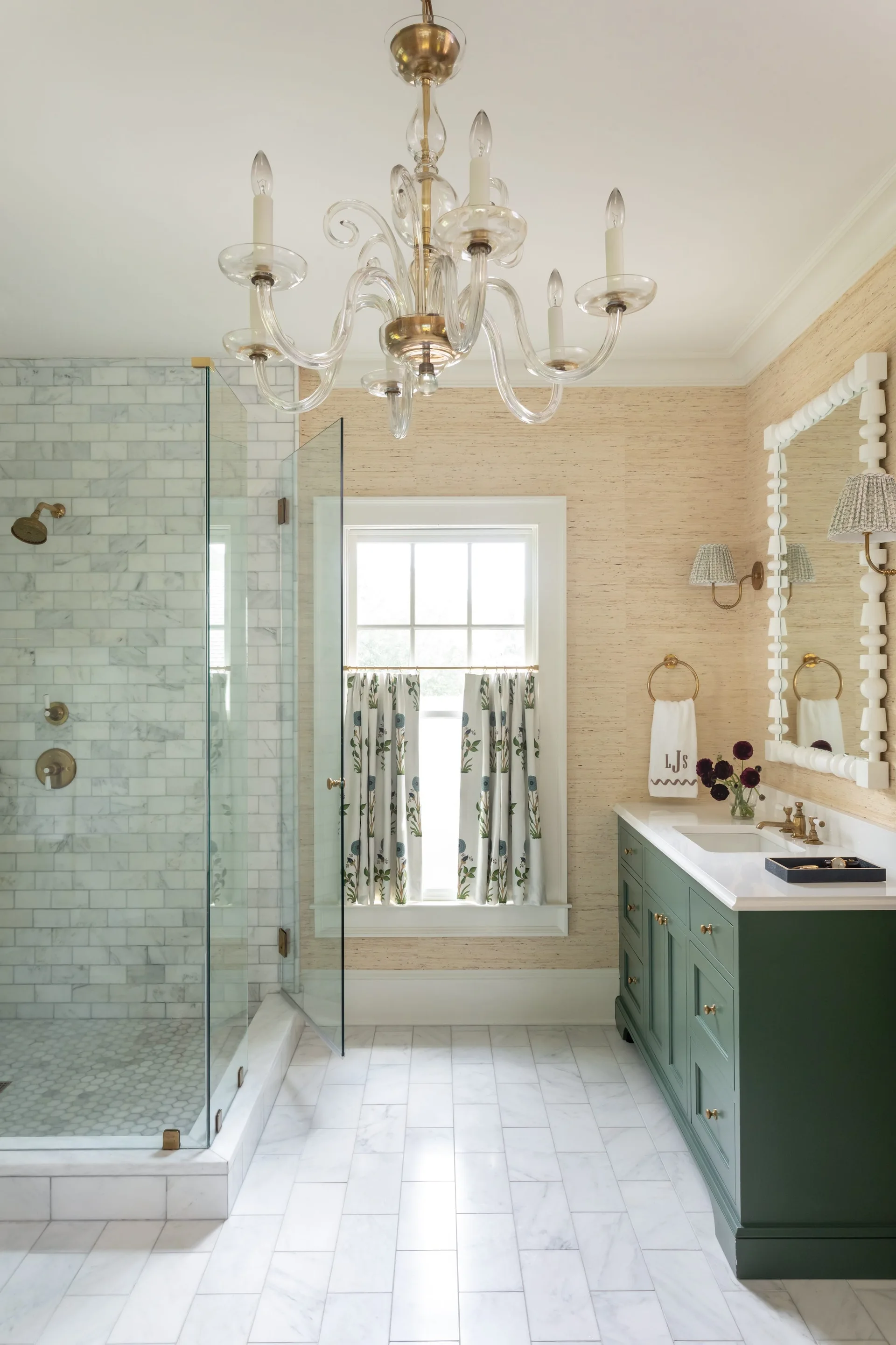 A traditional bathroom features a deep green vanity with brass hardware, a marble-tiled walk-in shower, and textured grasscloth wallpaper.
