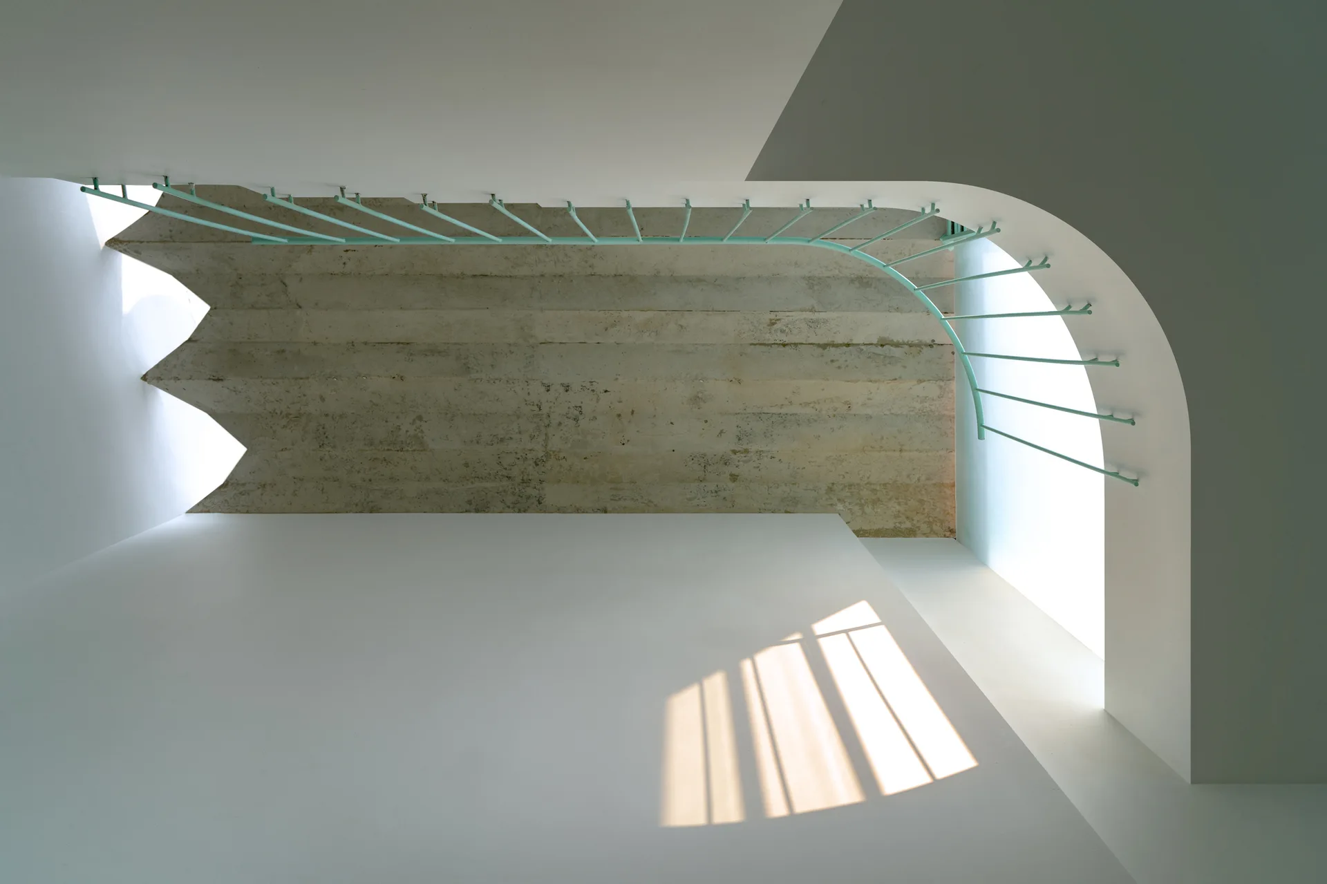 A minimalist white stairwell featuring a thin mint green metal railing against an exposed concrete ceiling and clean white walls.
