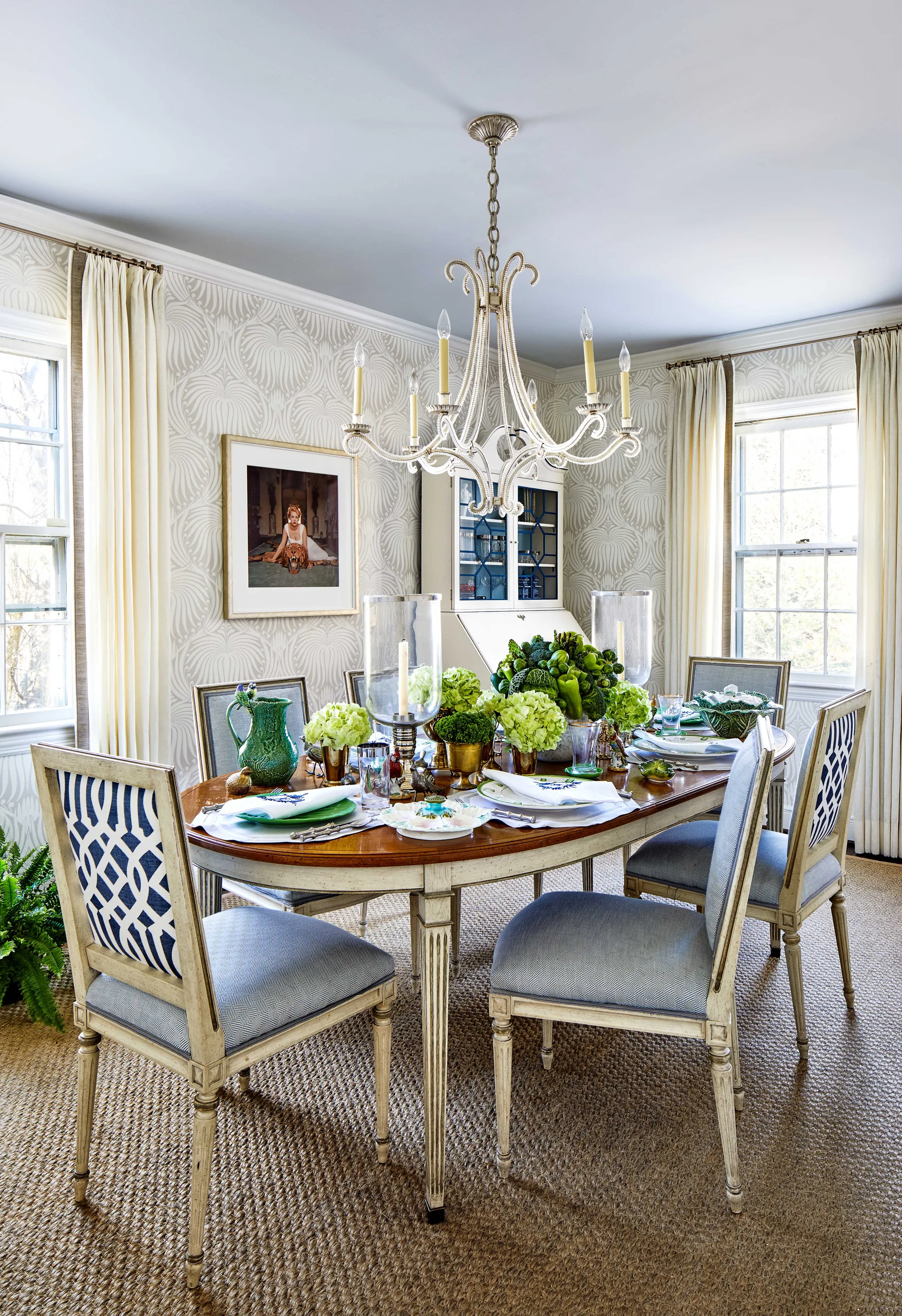 A formal dining room features an oval wooden table set with green decor, surrounded by blue upholstered chairs under an ornate white chandelier.