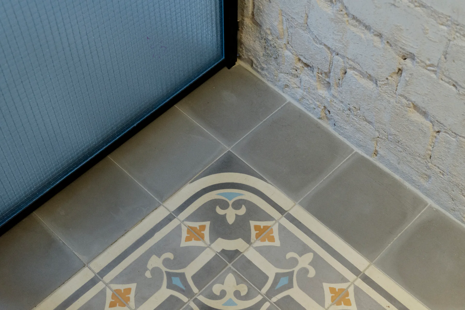 A close-up view of a grey tiled floor featuring a decorative patterned inlay beside a painted white brick wall and a framed glass partition.