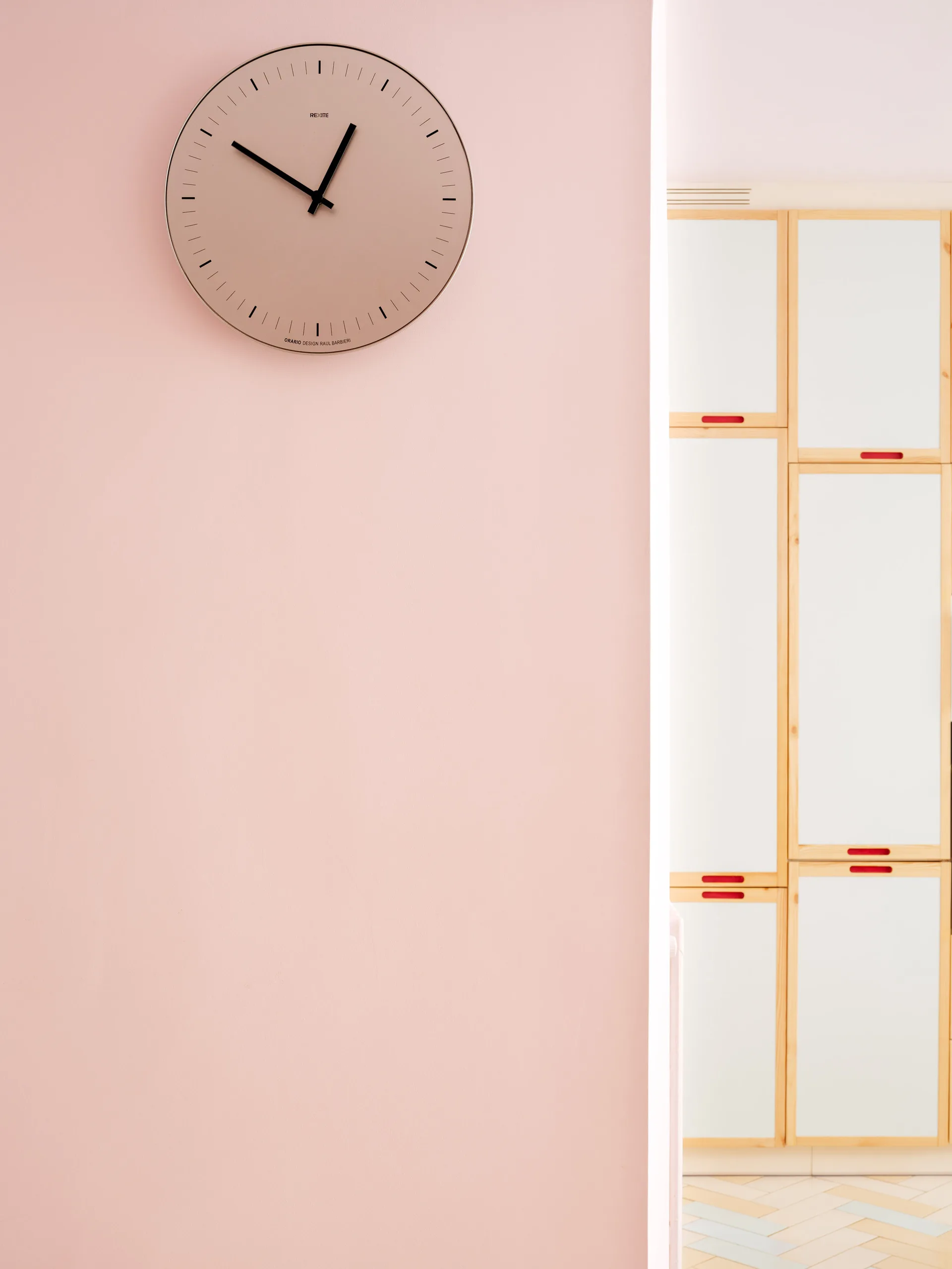 A minimalist round wall clock hangs on a soft pink wall next to white cabinetry with light wood trim and red handles.