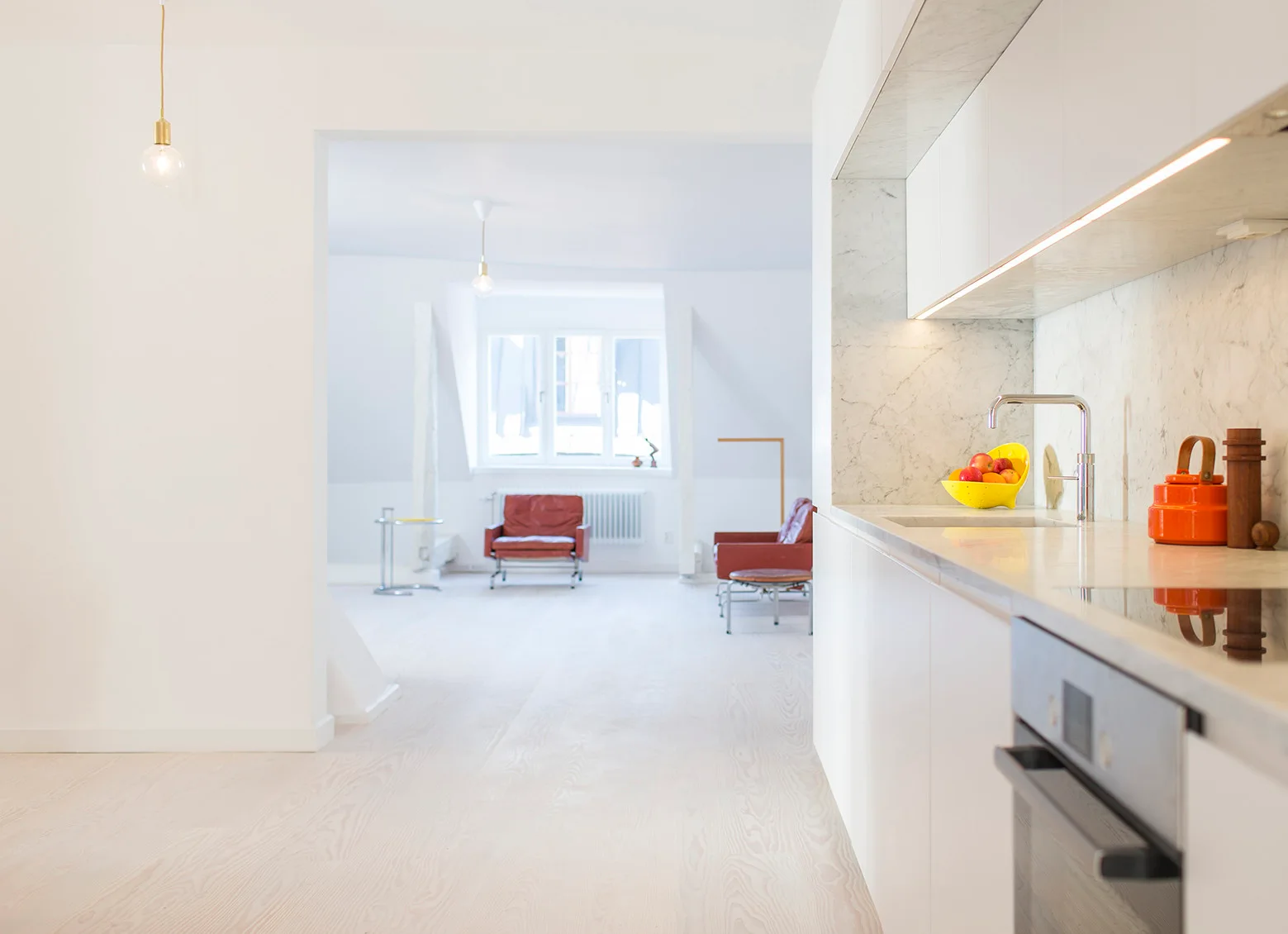 A minimalist white kitchen featuring marble countertops, flat-front cabinets, and light wood floors that extend into an adjacent open living space.