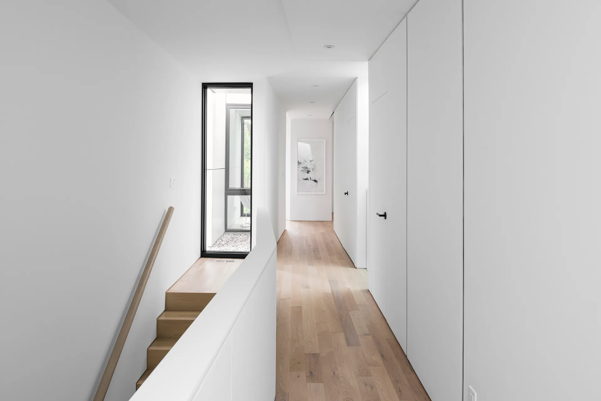 A bright white hallway with natural wood floors features a wooden staircase on the left and integrated flush white cabinet doors on the right.