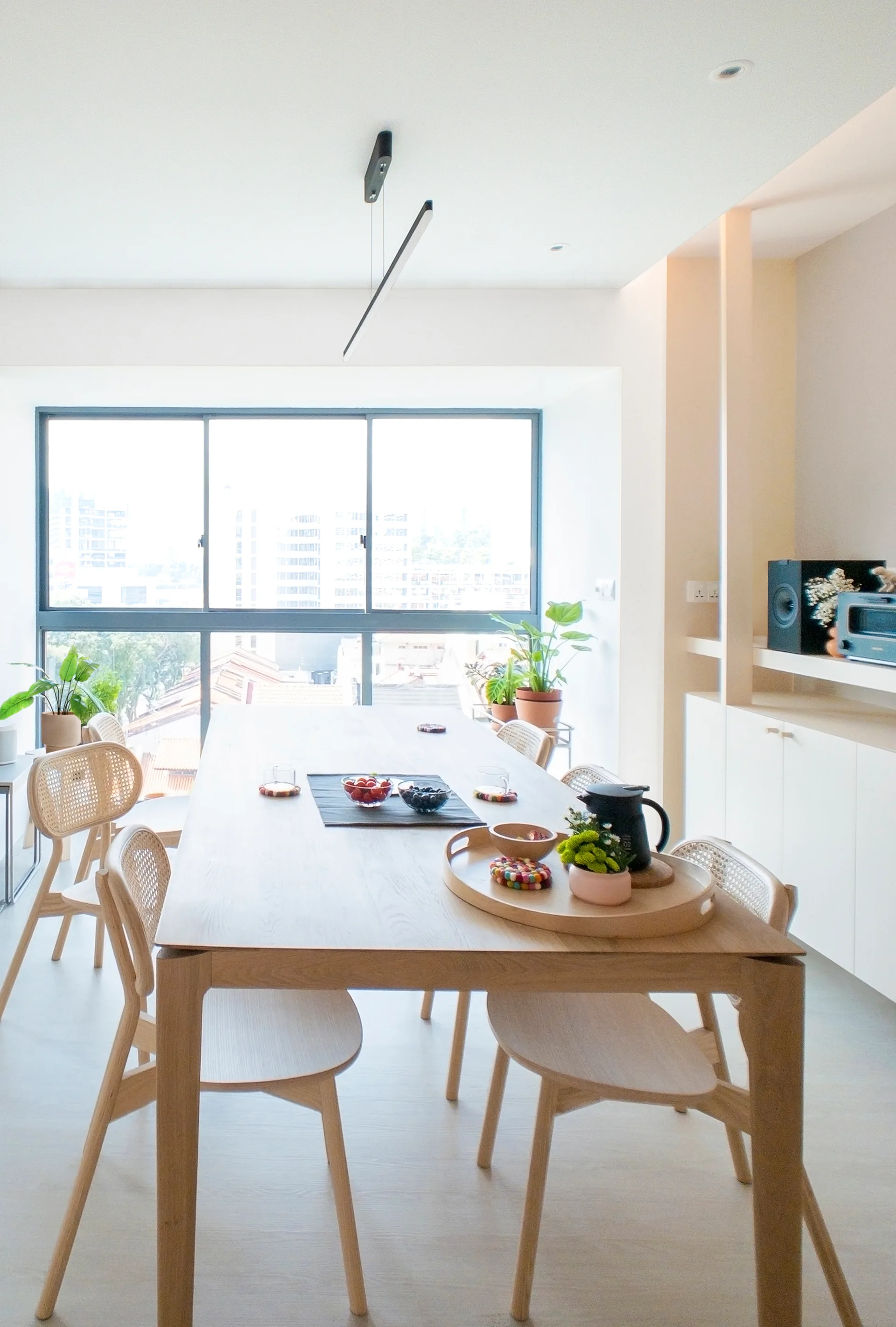 A bright dining room features a light wood dining table surrounded by wooden chairs with rattan backs near a large window.