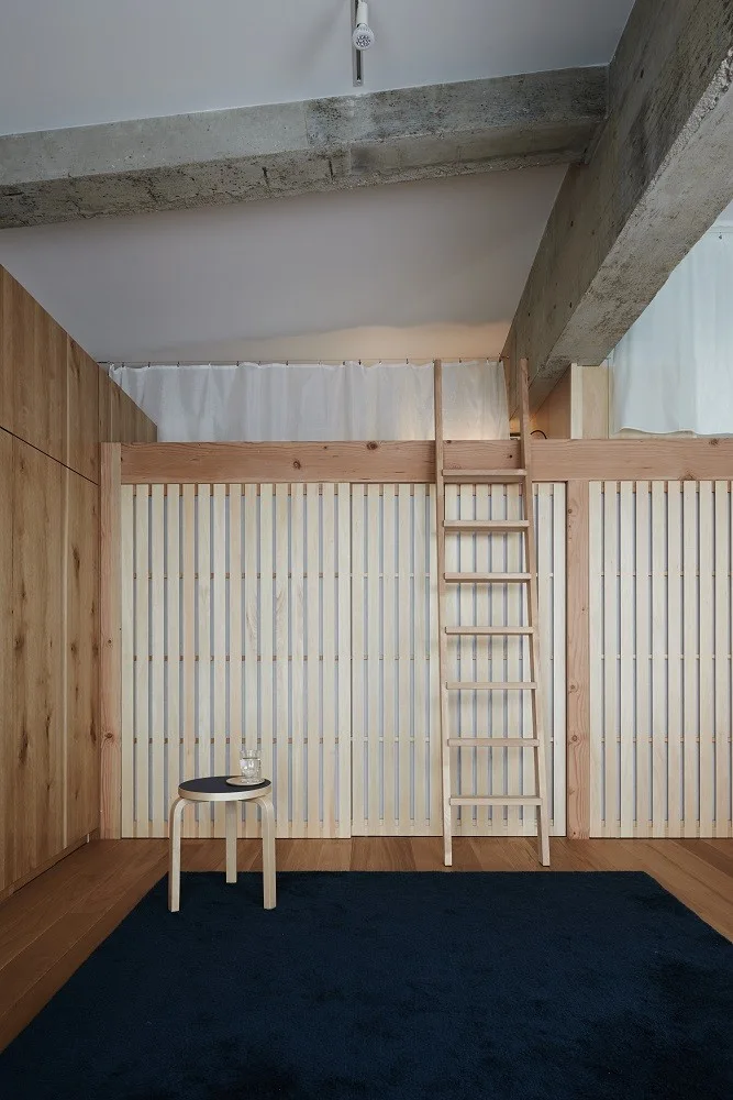 A minimalist loft bedroom features a light wood slatted partition wall, a matching leaning ladder, a small stool, and a navy blue area rug.