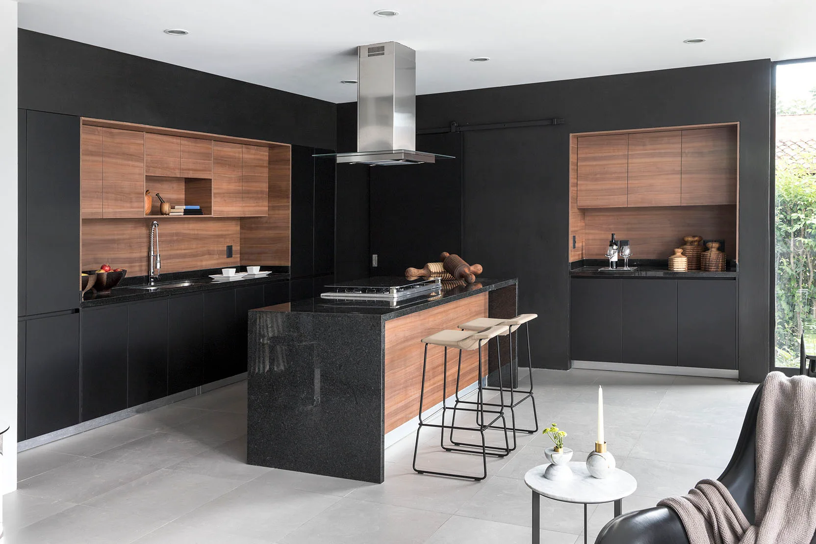 A modern kitchen featuring matte black wall cabinets with warm wood-toned niches, a black stone island with high stools, and light grey tile flooring.