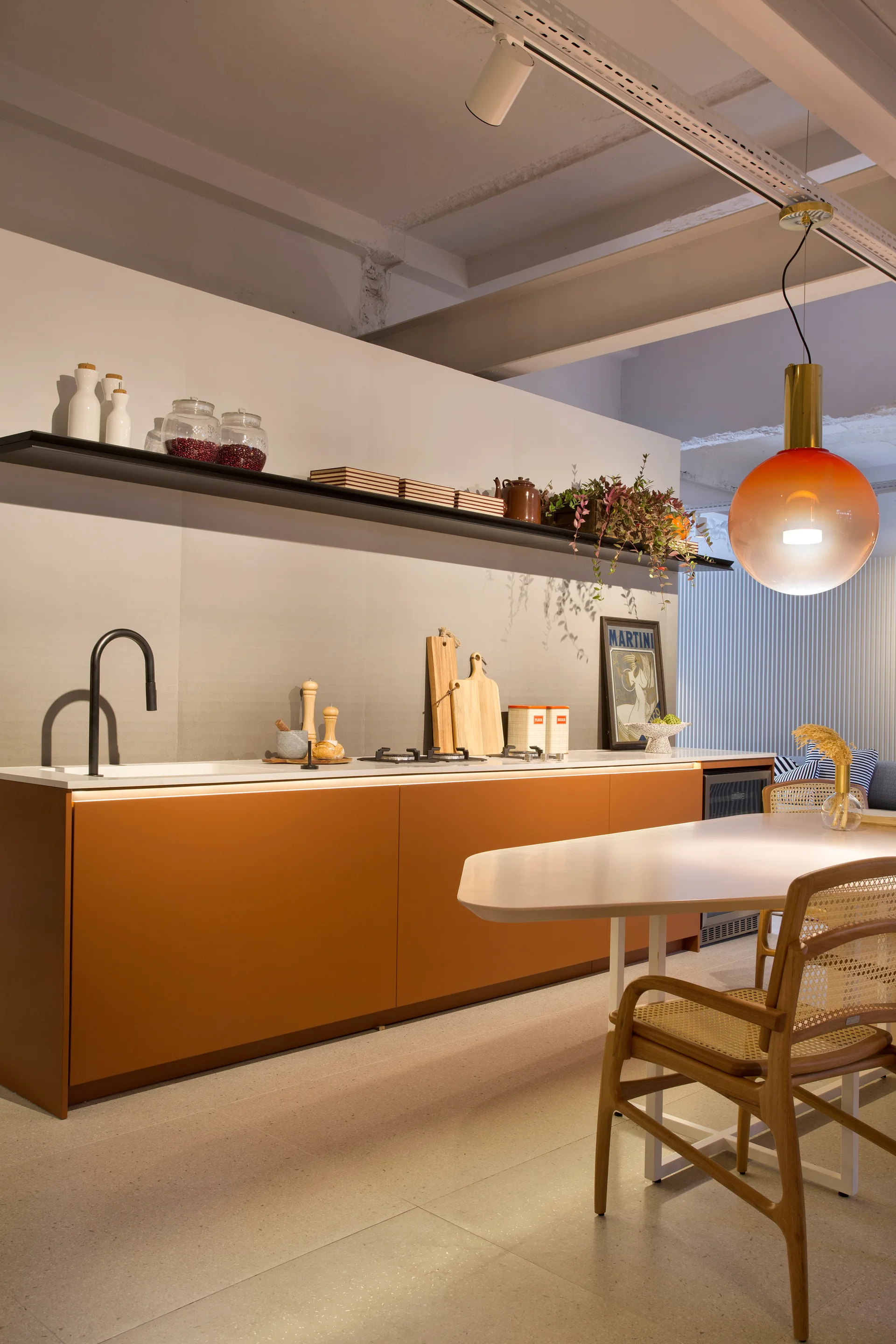 A modern kitchen features matte orange base cabinets under a white countertop, paired with a light-colored dining table and wooden cane-back chairs.