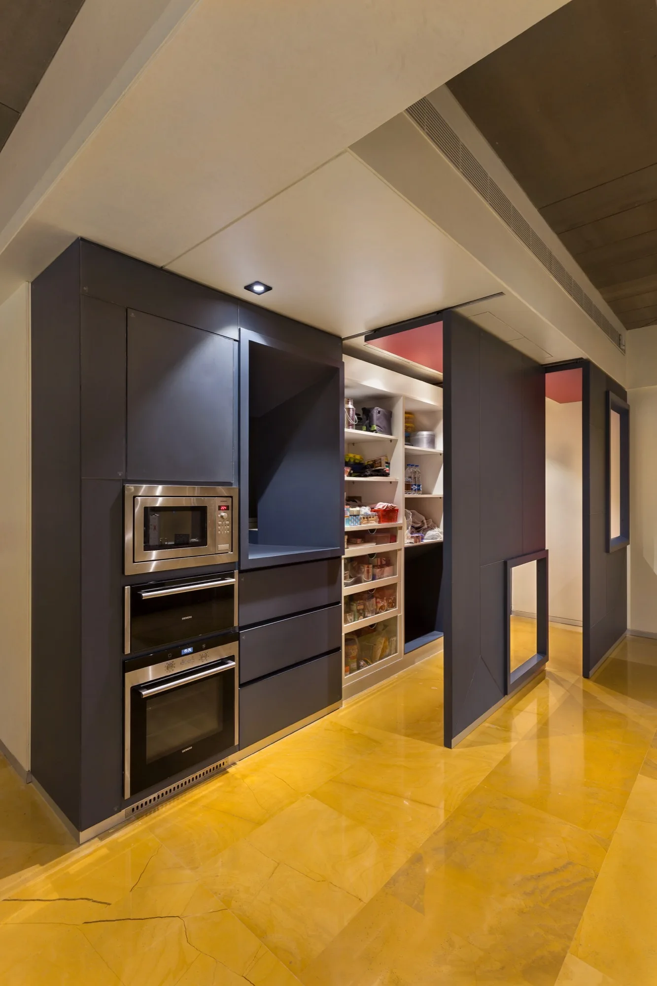 Charcoal gray kitchen cabinets with built-in ovens and a sliding panel that reveals a stocked pantry on yellow polished floors.